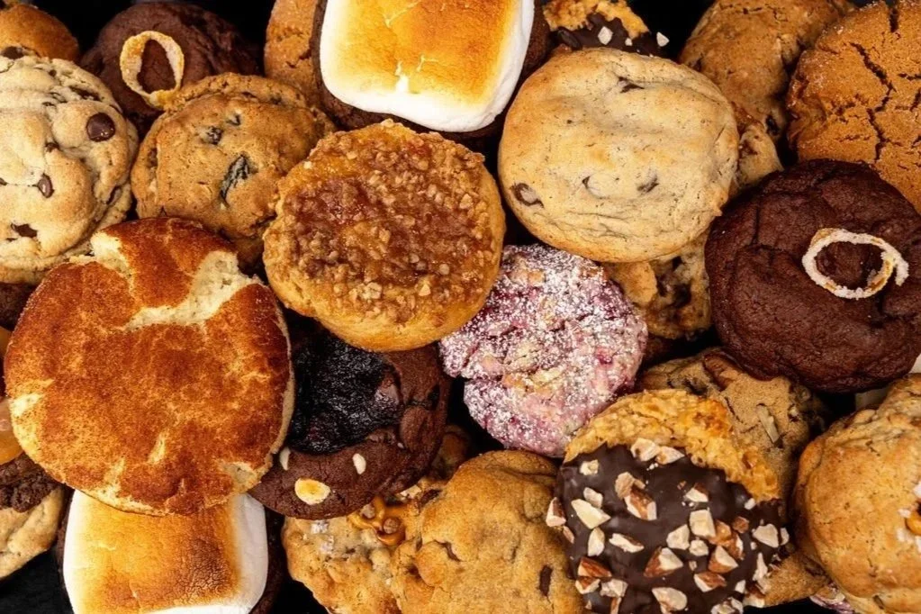 Assorted freshly baked cookies including chocolate chips, oatmeal, sugar cookies, and cookies with nuts and icing.