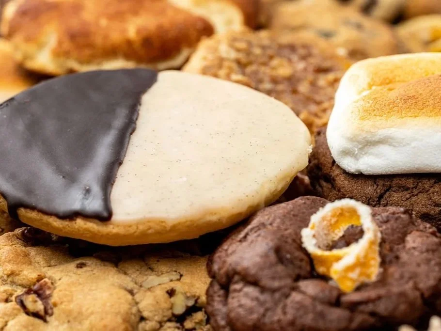 assortment of cookies, including a half-dipped chocolate cookie, a white frosted cookie, and a chocolate cookie with an orange peel decoration, with more cookies in the background