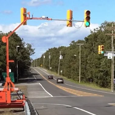A traffic light hanging over a two-lane road with cars, surrounded by trees and blue sky.