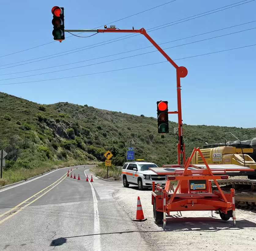 Traffic control light system with red lights flashing, set up on a road with construction cones and a parked vehicle. Construction equipment and a call box are visible near a hilly landscape.