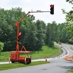 Traffic light mounted on an orange utility vehicle on the side of a curved road with trees and construction cones.