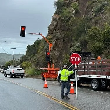 A worker in a high-visibility jacket holding a stop sign at a roadside construction zone with traffic cones, a SQ3TS portable stoplight from Tysen Company, and a truck on a curved road next to rocky terrain and greenery.