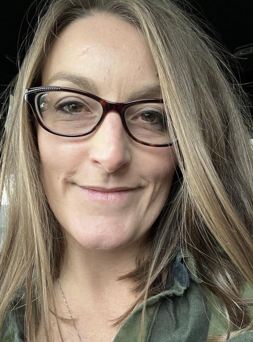 A close-up photo of a smiling woman with light skin, wearing rectangular tortoiseshell glasses and a camouflage jacket, with long blonde hair.