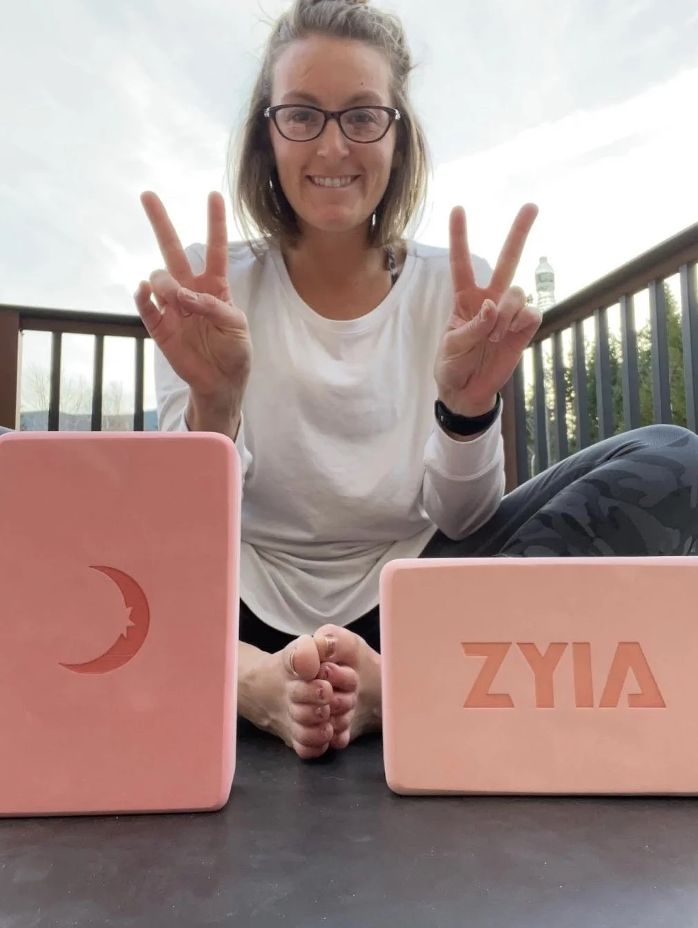 Woman sitting outdoors on a deck, smiling, making peace signs with both hands. She has glasses, blond hair, and is wearing a white long sleeve shirt. Two pink laptops are in front of her, one with a moon and star design and the other with the word "Aiya" cut out. A water bottle is visible on the railing.