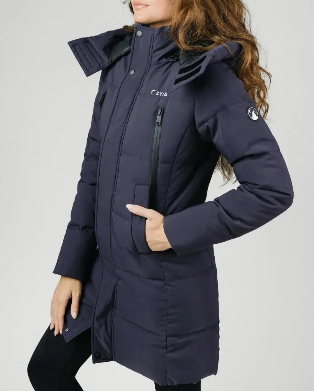 A woman in a dark blue winter coat with a hood, standing with one hand in her pocket.