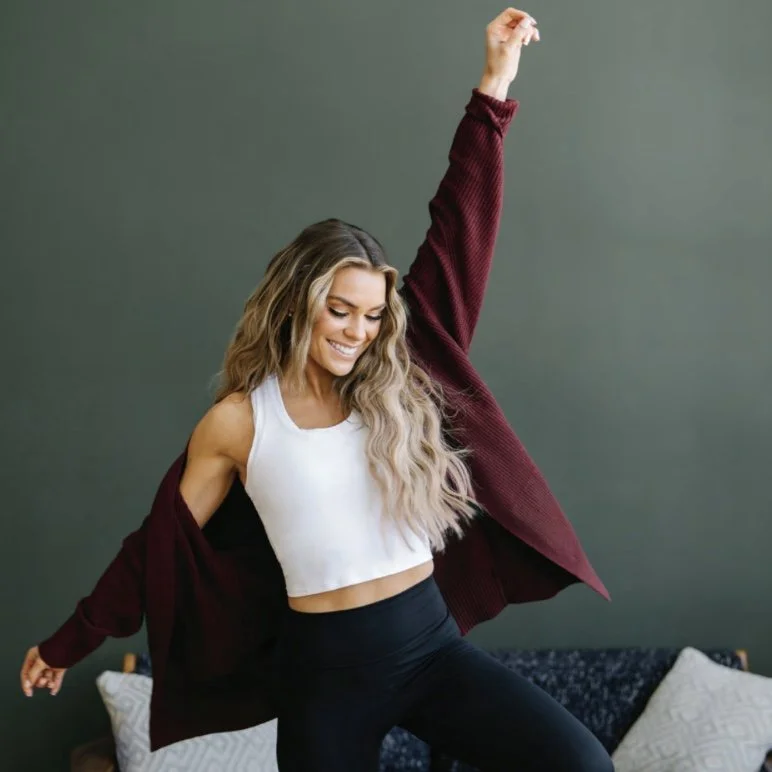 A woman with long wavy hair dancing joyfully indoors, wearing a white crop top, black leggings, and a maroon sweater draped off her shoulders, against a green wall.