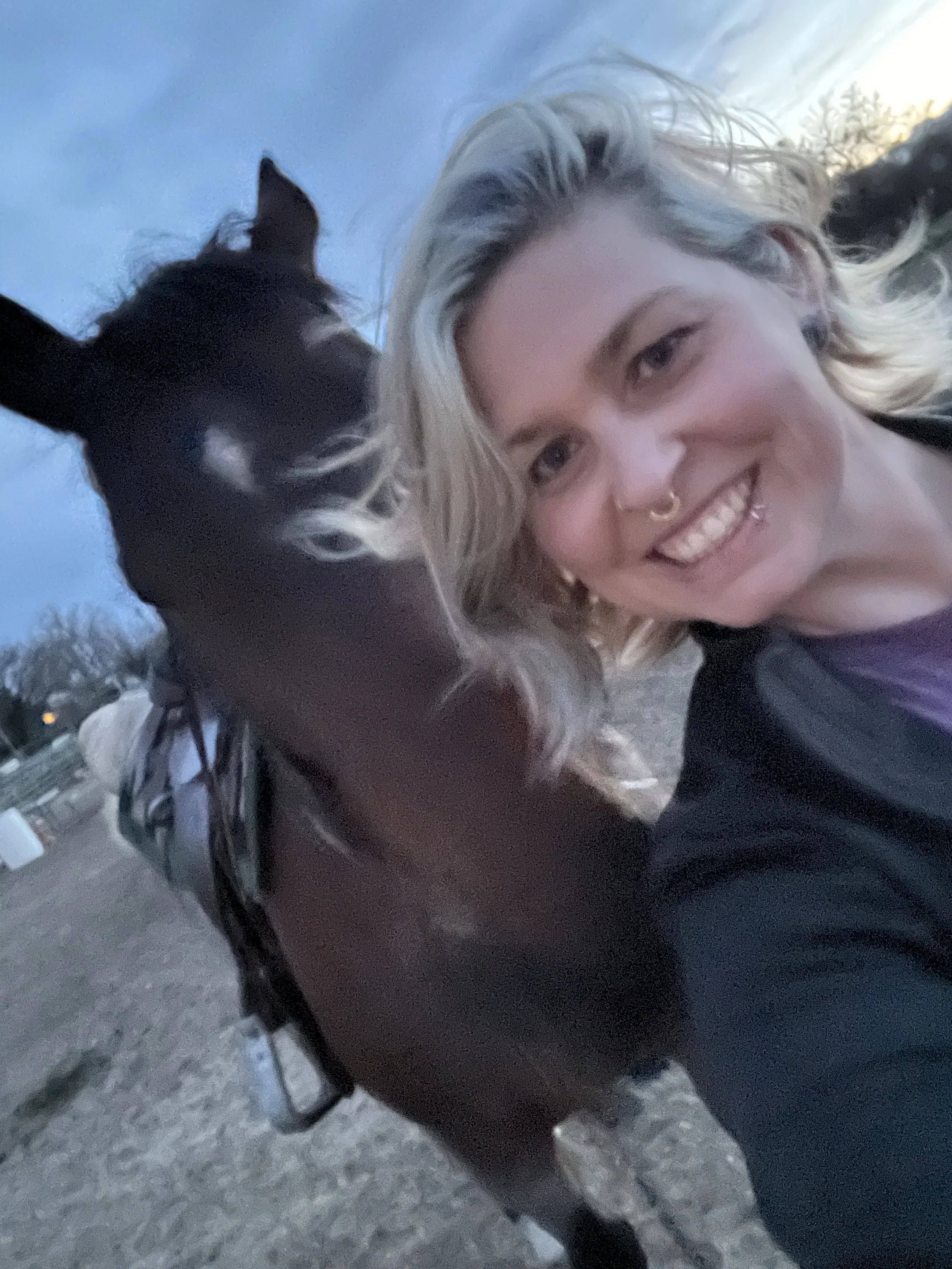 Young woman with blonde hair smiling next to a dark brown horse during twilight in an outdoor setting.