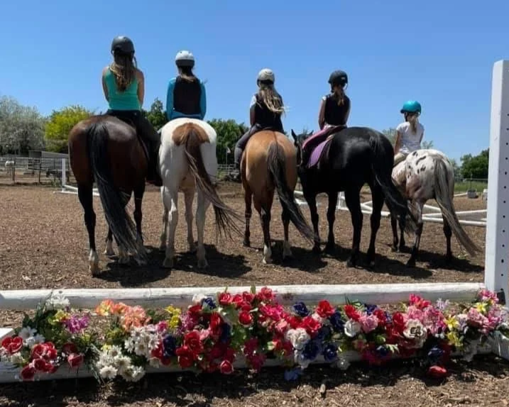 Six people, all wearing helmets, are riding horses in an outdoor riding arena on a sunny day. A colorful flower arrangement is in the foreground.