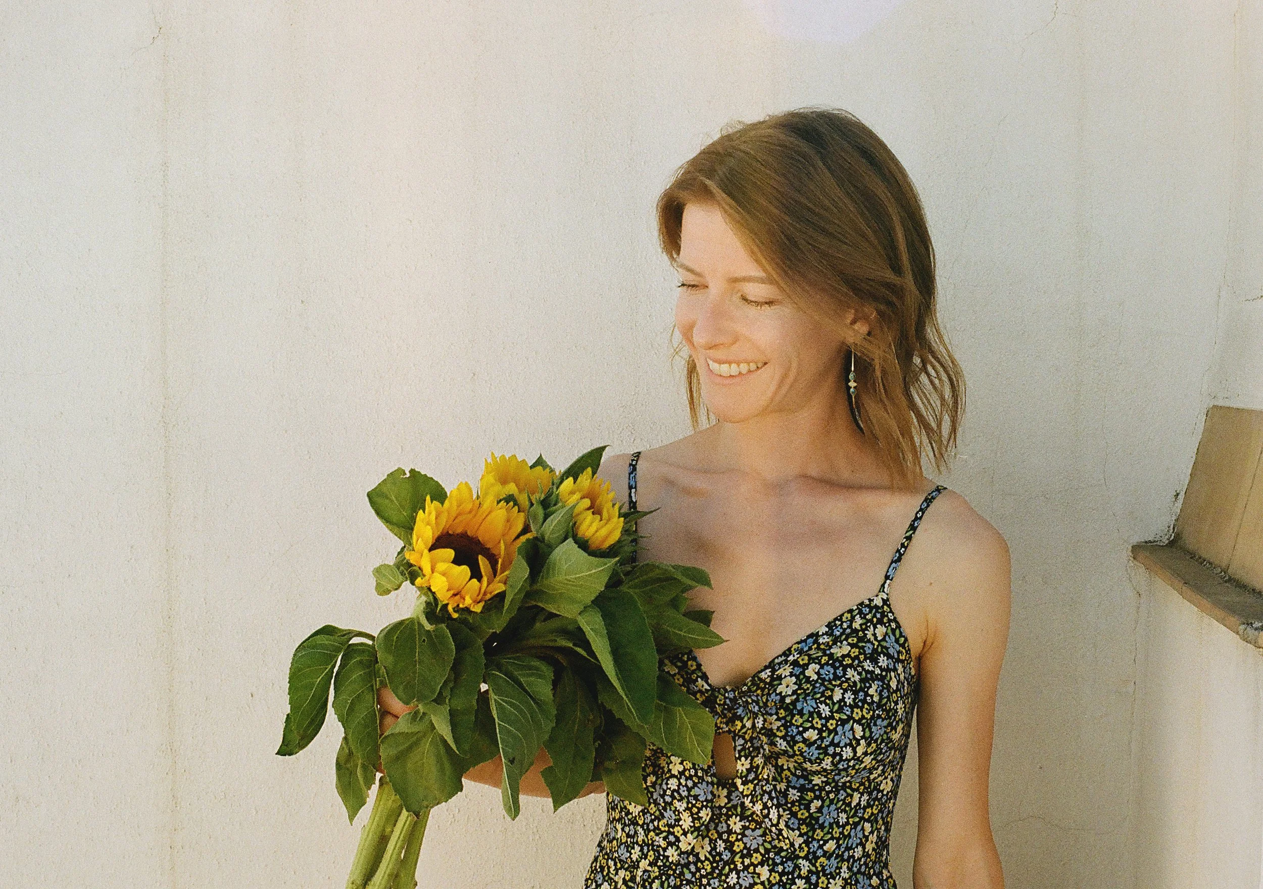 A woman with shoulder-length brown hair smiling while holding a bouquet of sunflowers and greenery, standing against a light-colored wall.