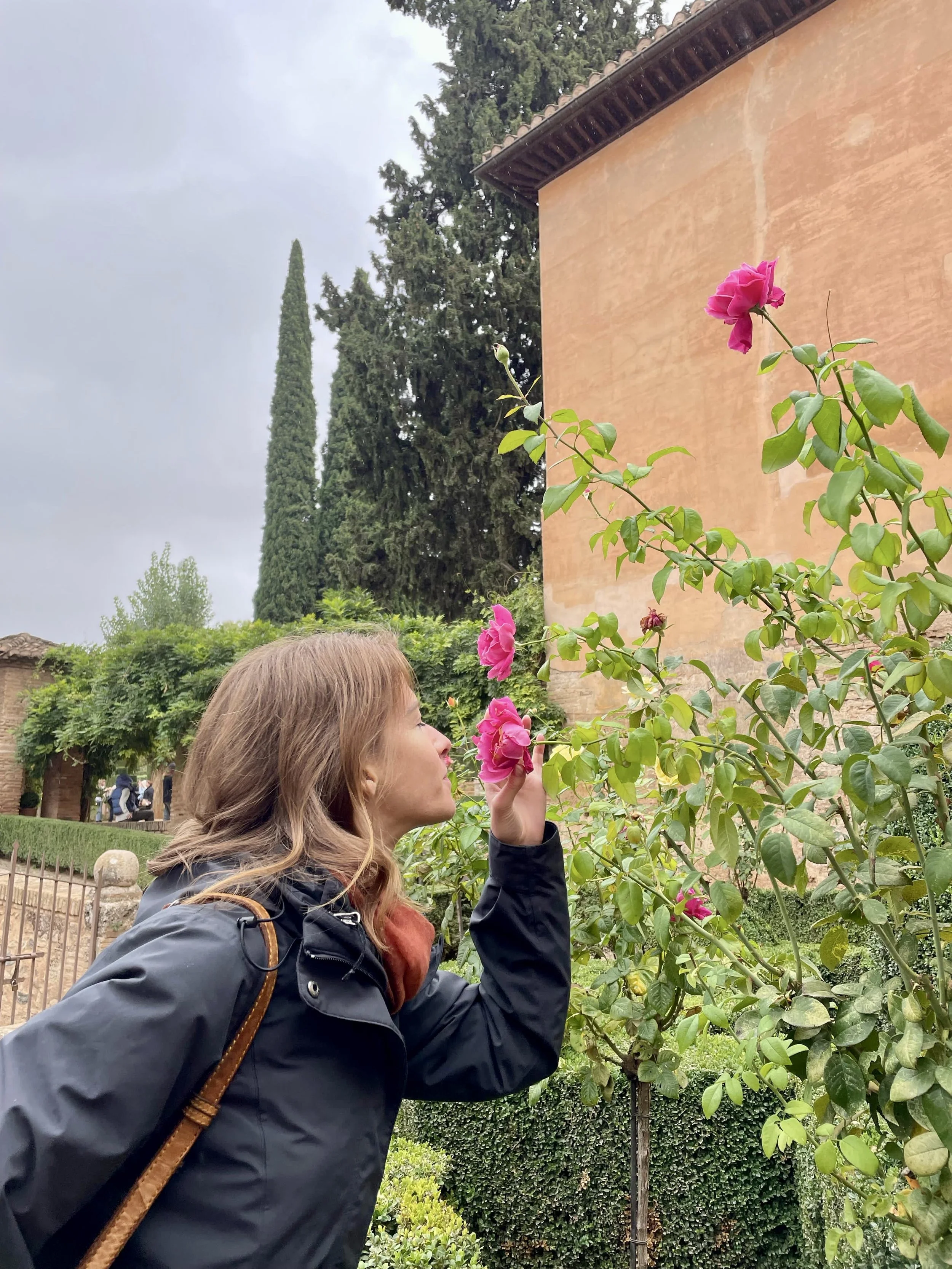 A woman smelling a pink rose on a bush near a peach-colored building, with greenery and tall cypress trees in the background on a cloudy day.