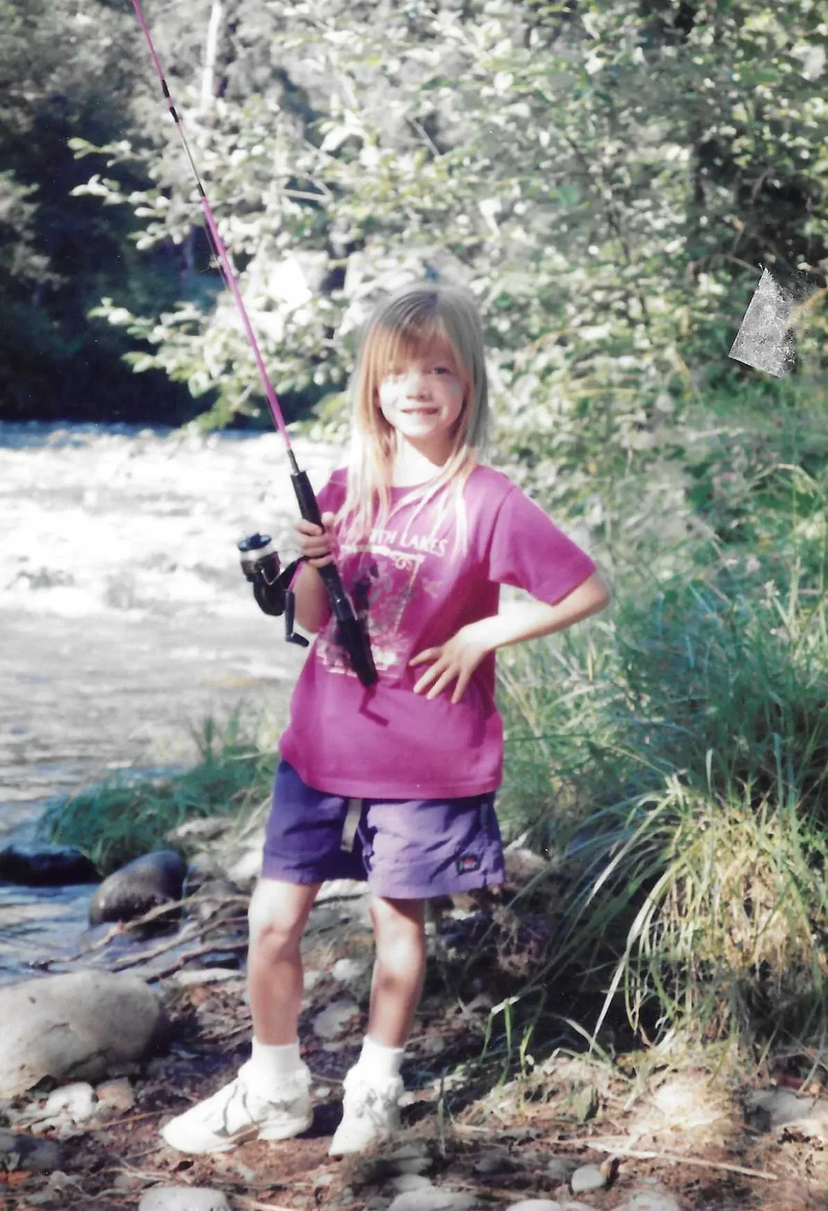 Young girl in purple shirt and shorts holding a fishing rod, standing by a river with trees in the background.