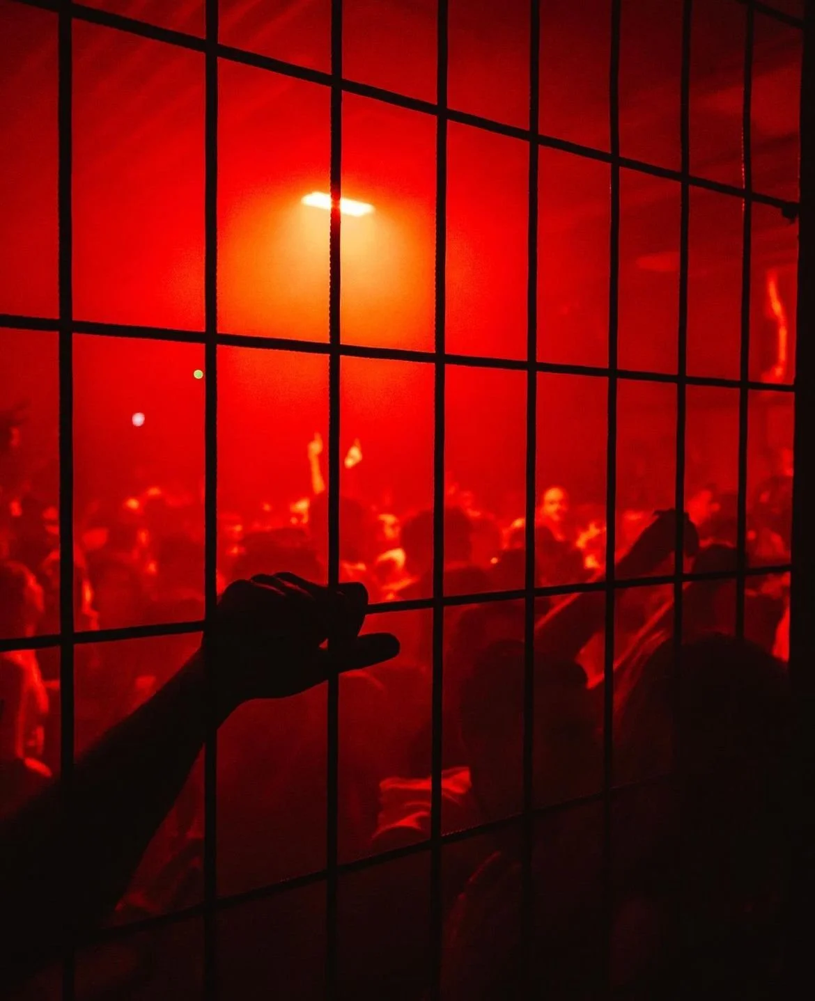 People behind a wire fence at a concert with red lighting.