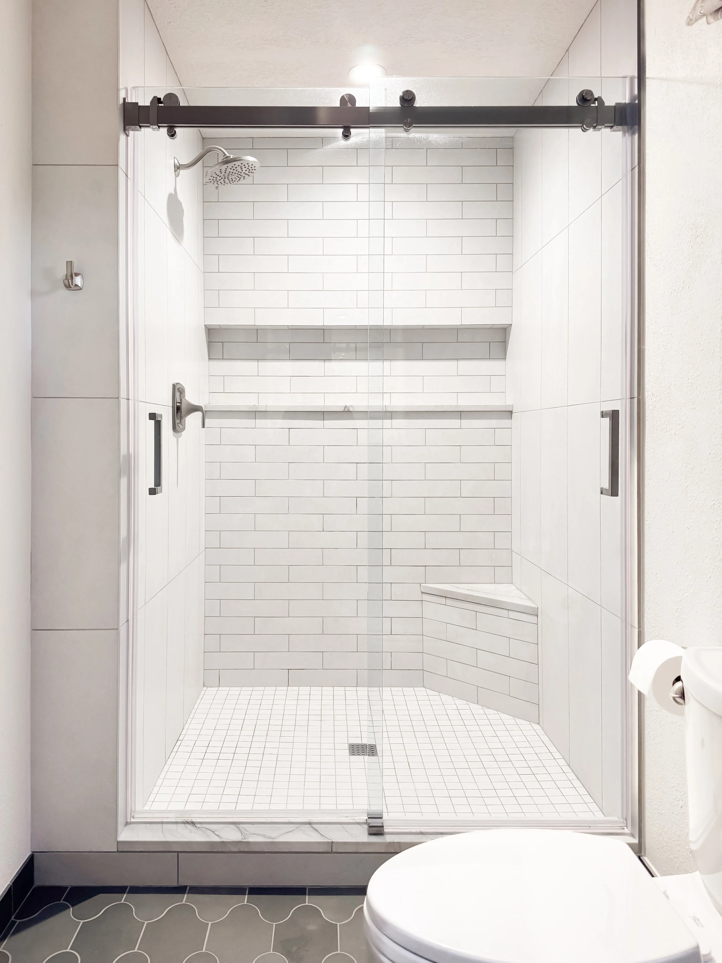 A modern bathroom with a walk-in glass shower featuring white subway tile walls, a built-in shelf, a chrome showerhead, and a sliding door. A white toilet is partially visible in the foreground, with a black tiled floor and white walls.