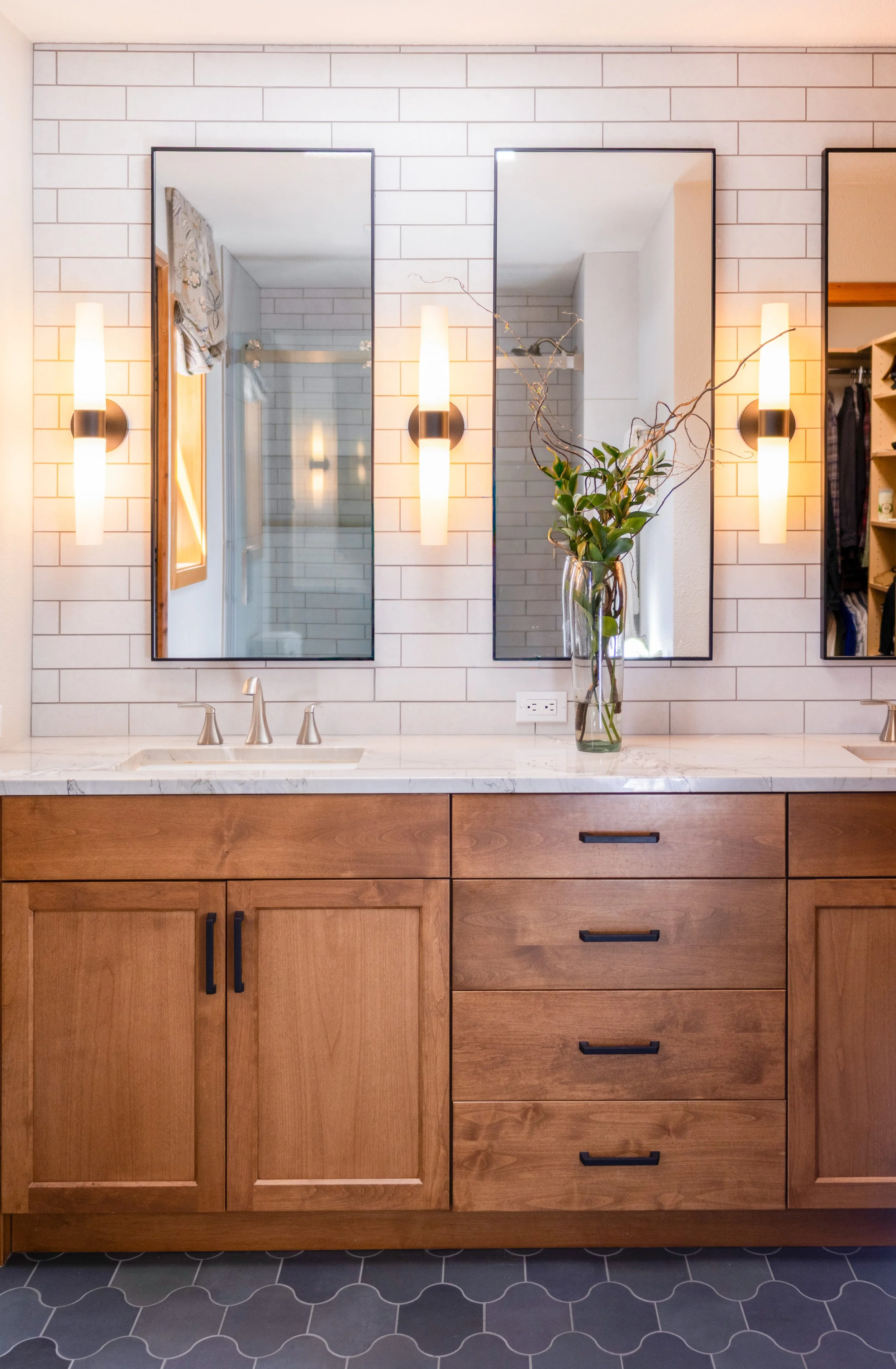 Bathroom vanity with a white marble countertop, wooden cabinet, and three drawers. Two vertical wall mirrors with black frames. Two wall-mounted light fixtures between the mirrors. A glass vase with green leaves and twigs on the countertop. Hexagonal dark gray floor tiles.