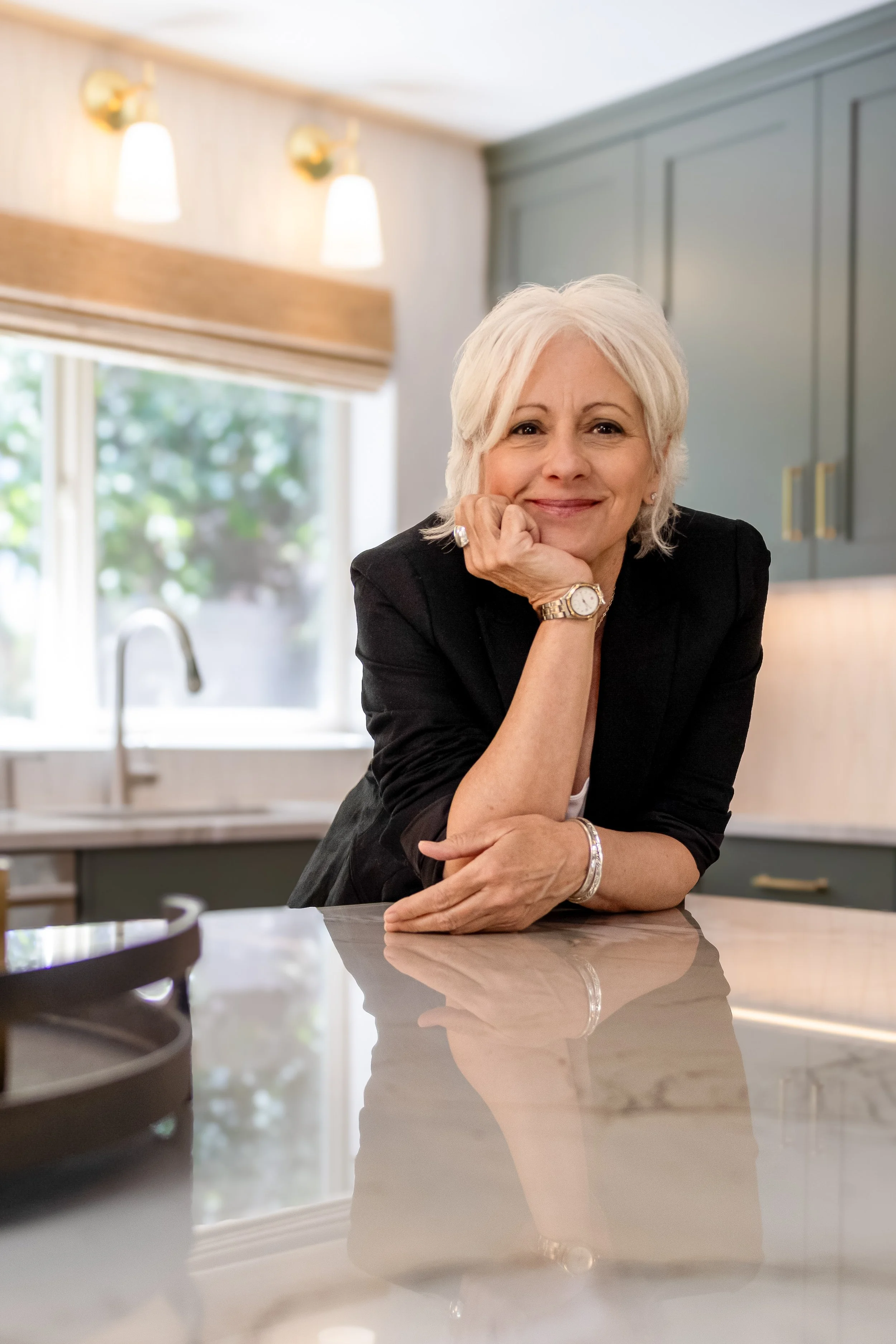 A woman with short white hair smiling and resting her chin on her hand in a kitchen with a window, grey cabinets, and modern lighting.