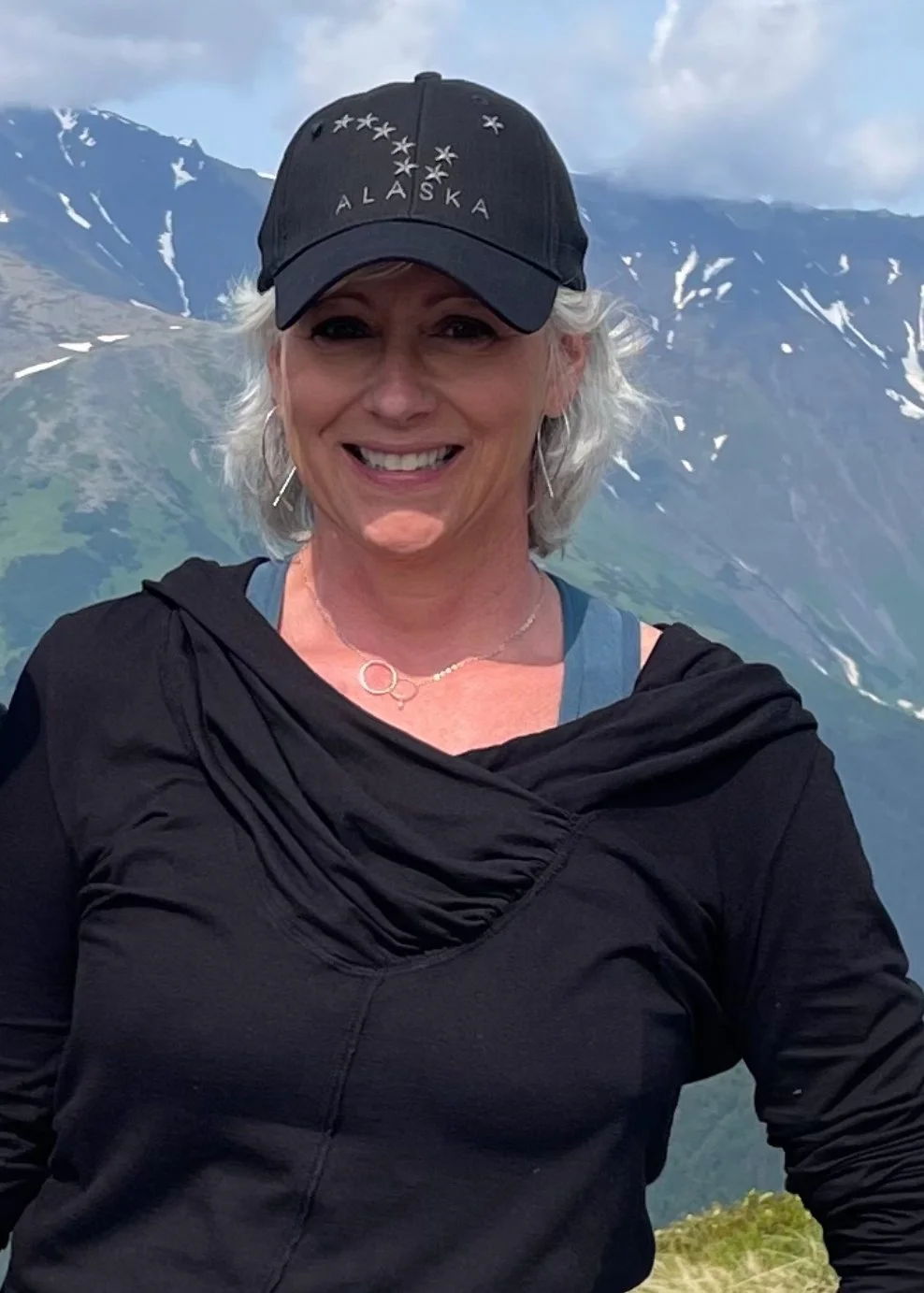 Smiling woman with short gray hair wearing a black hat with 'Alaska' written on it, black jacket, and blue top, standing outdoors with mountains and snow in the background.