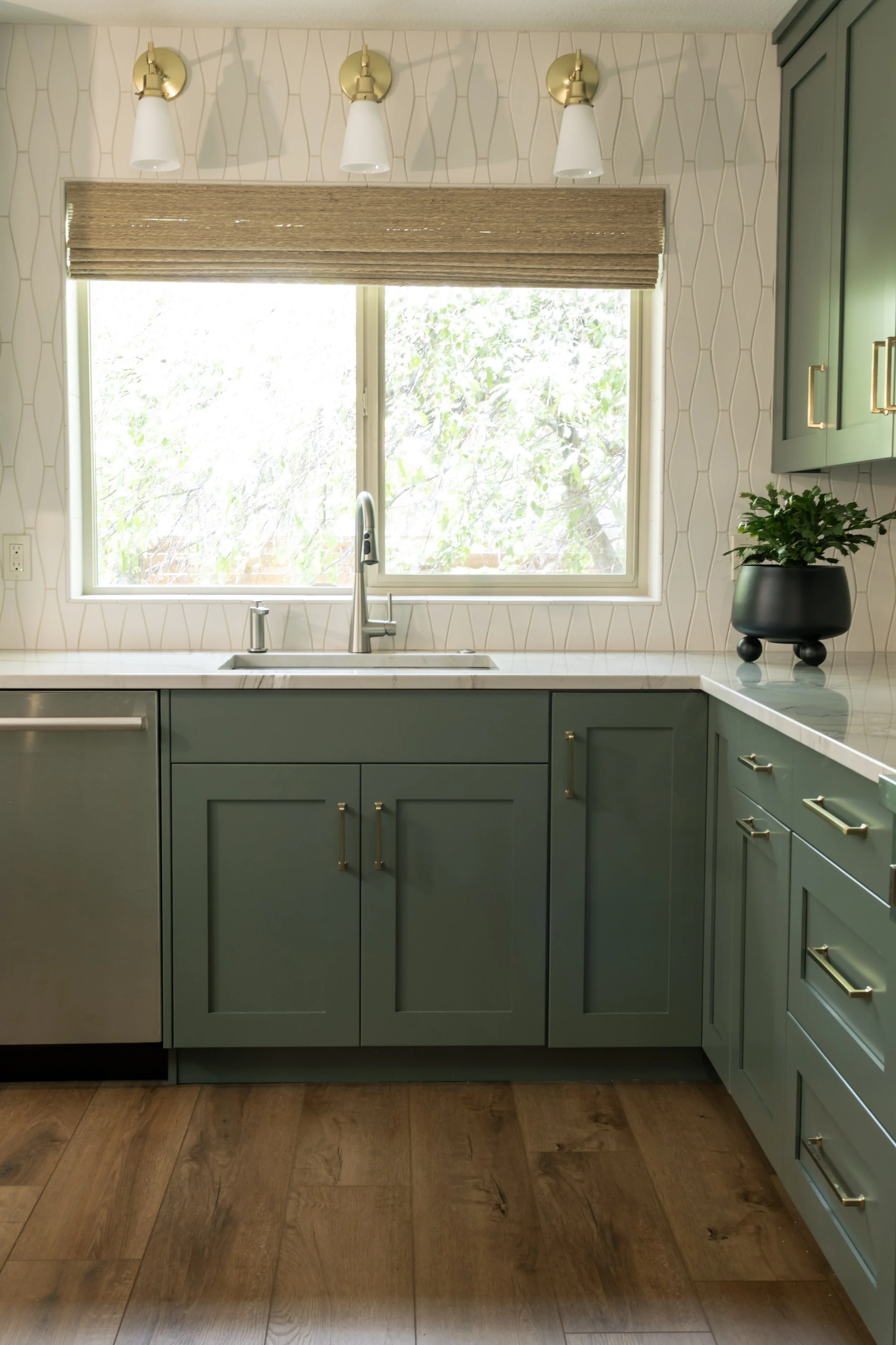 A kitchen with teal cabinets, a white marble countertop, a window above the sink, and a potted plant on the counter. There are three white and gold wall-mounted light fixtures above the window and a beige woven shade covering part of the window.