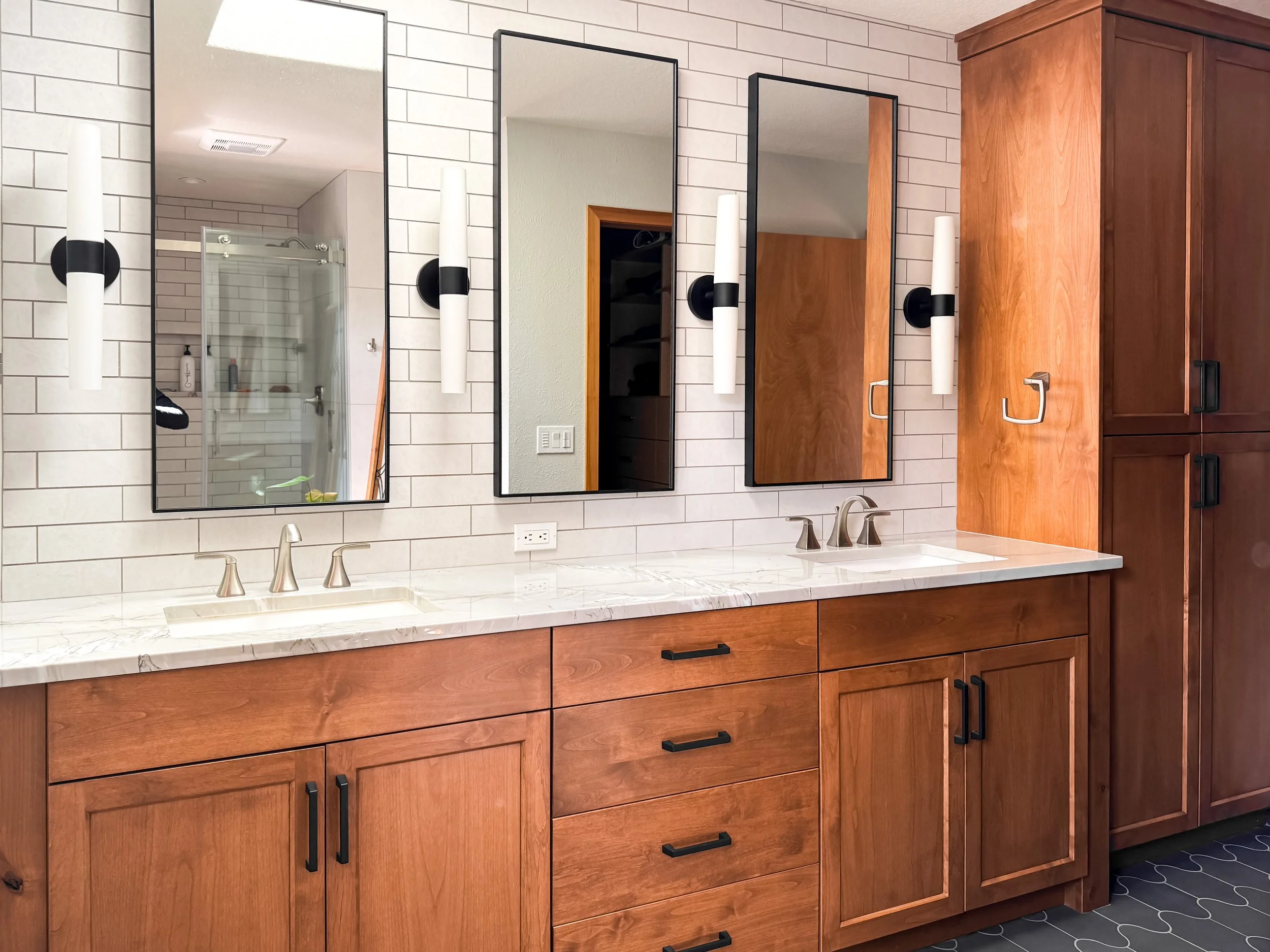 A bathroom vanity with a white marble countertop, two undermount sinks, and modern brushed nickel faucets. Above the vanity are three rectangular mirrors with black frames, flanked by black and white wall sconces. Wooden cabinets with black handles are below the countertop and to the right, providing storage.