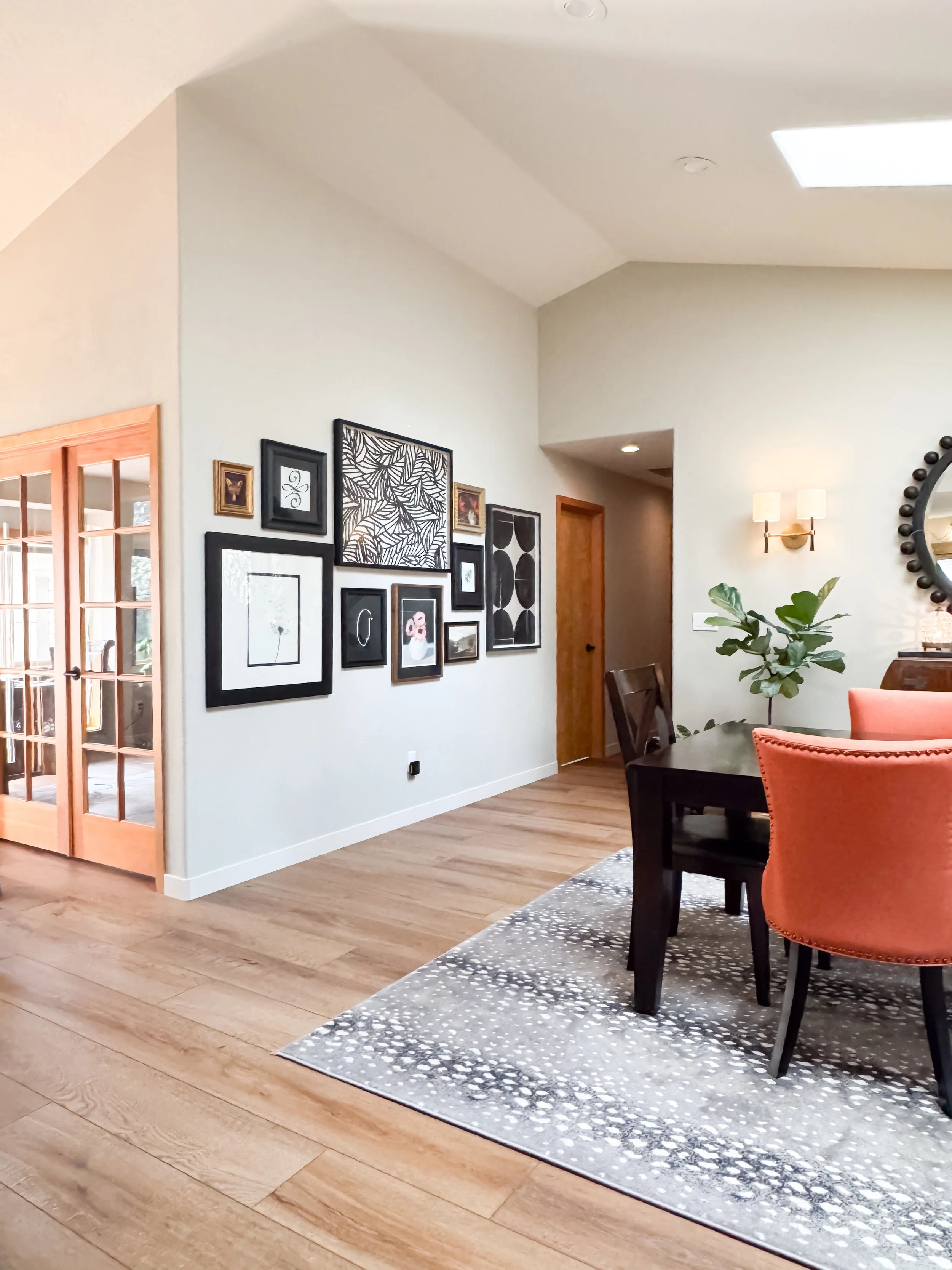 A dining room with wooden flooring, black dining table, orange upholstered chairs, a patterned area rug, wall collage, green potted plant, wall-mounted light fixtures, and a mirror.
