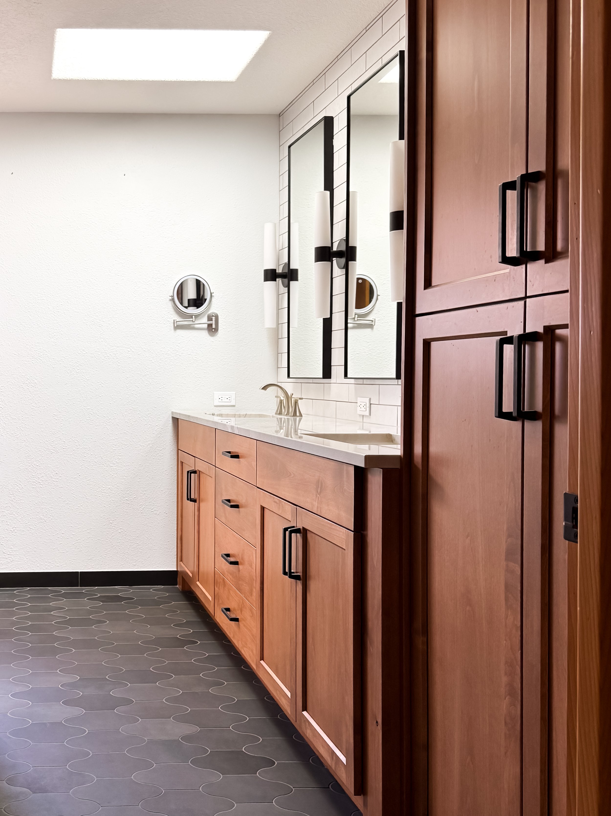 Interior view of a bathroom with dark gray hexagonal floor tiles, wooden cabinets with black handles, a white countertop with a built-in sink, two black-framed mirrors, wall-mounted light fixtures, a white textured wall, and a white ceiling with a re