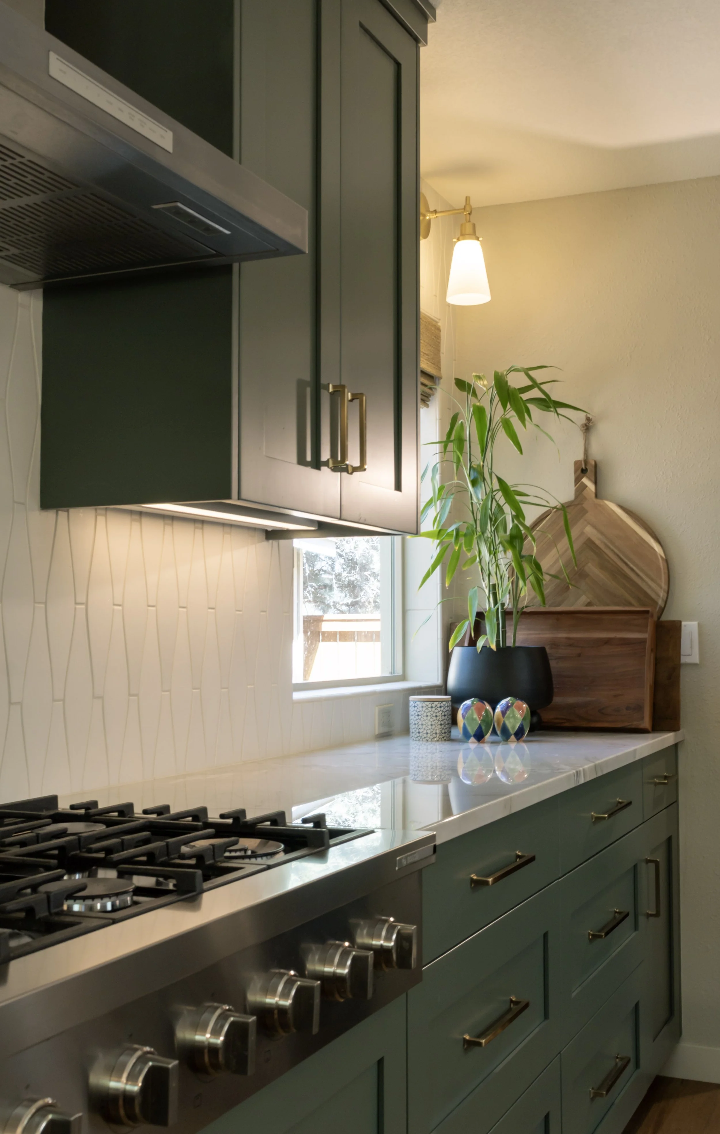A modern kitchen with green cabinets, a white marble countertop, a gas stove, decorative vases, a potted plant, and a window letting in natural light.