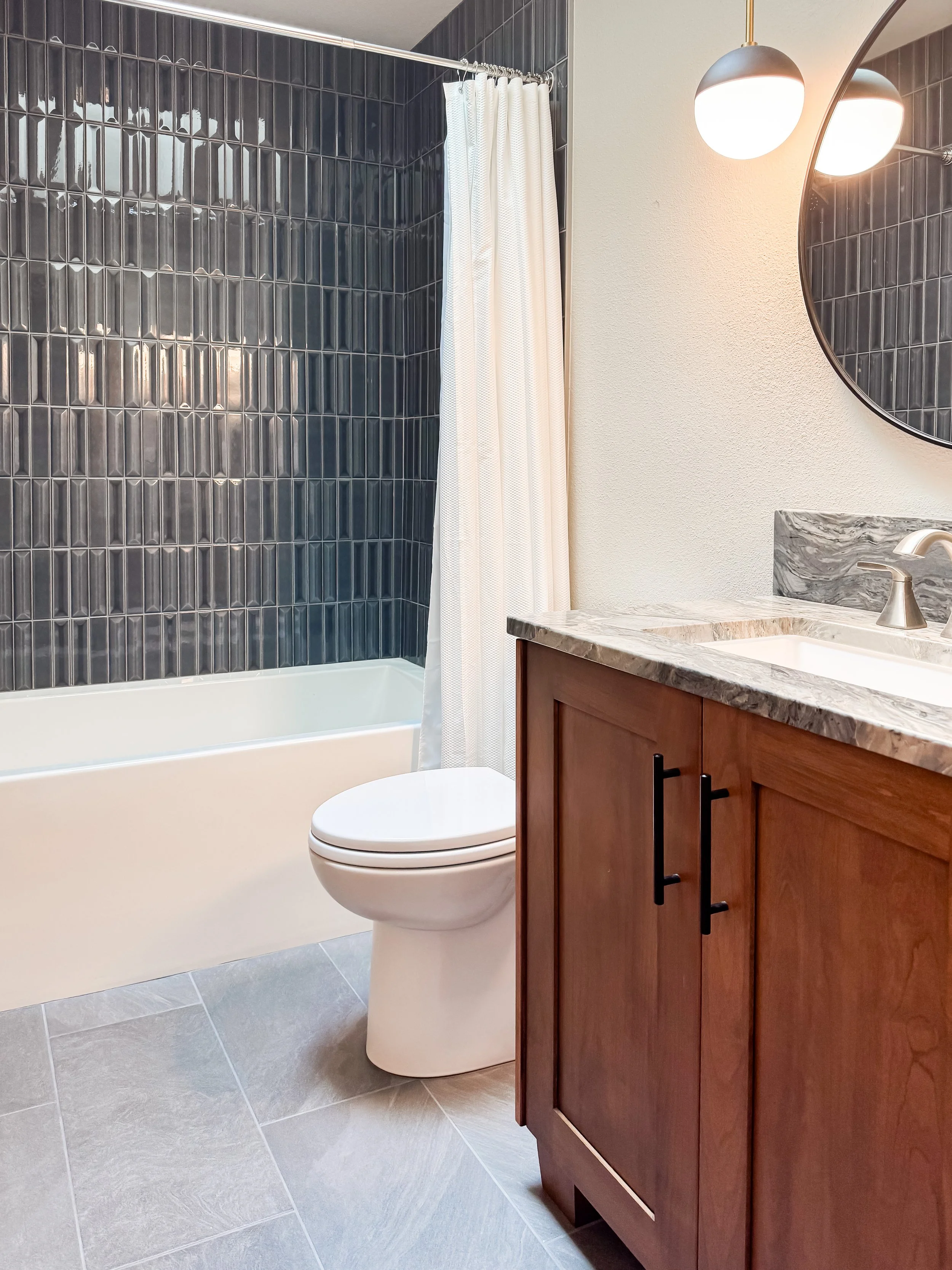 Bathroom with black tiled shower, white curtain, wooden vanity with marble countertop, round mirror, wall-mounted light, and toilet.