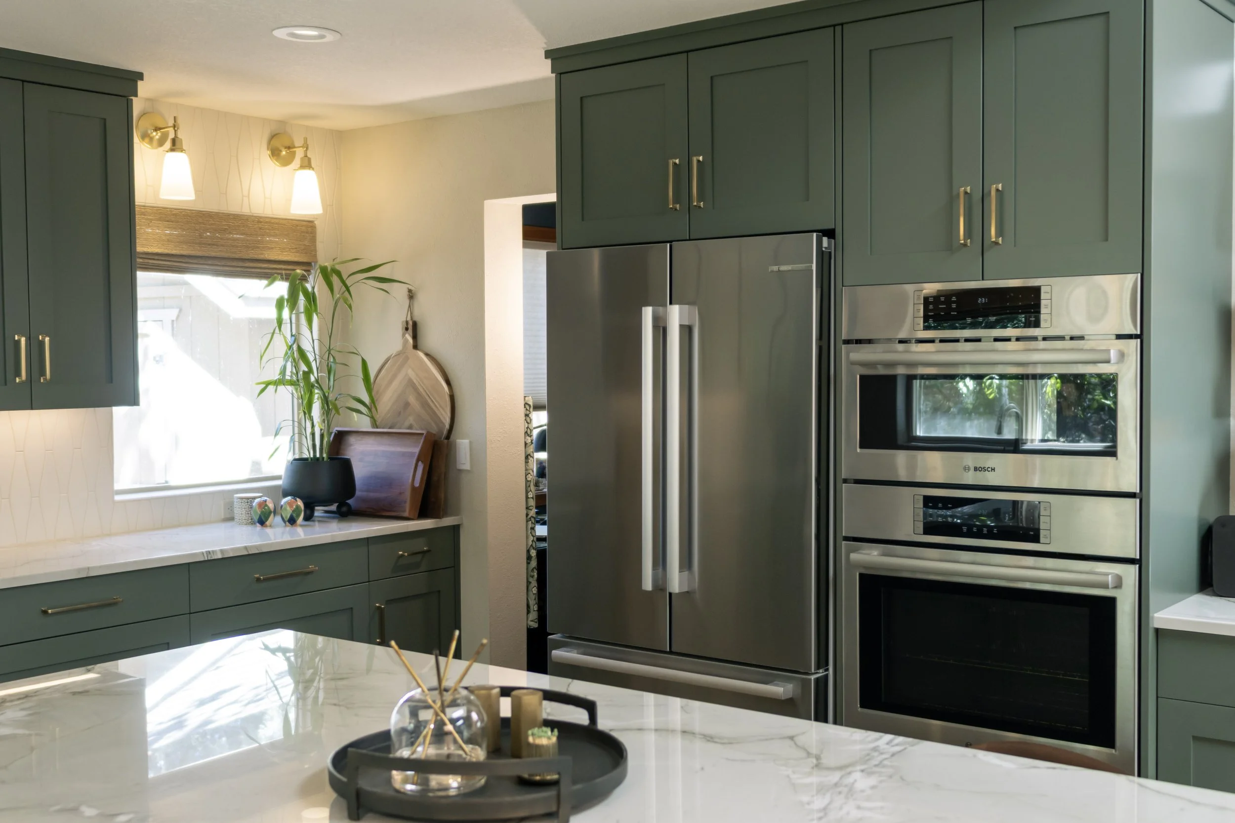 Green kitchen cabinets with gold handles, stainless steel refrigerator, double oven, window with blinds, plant on the counter, decorative tray and candles on the marble kitchen island.