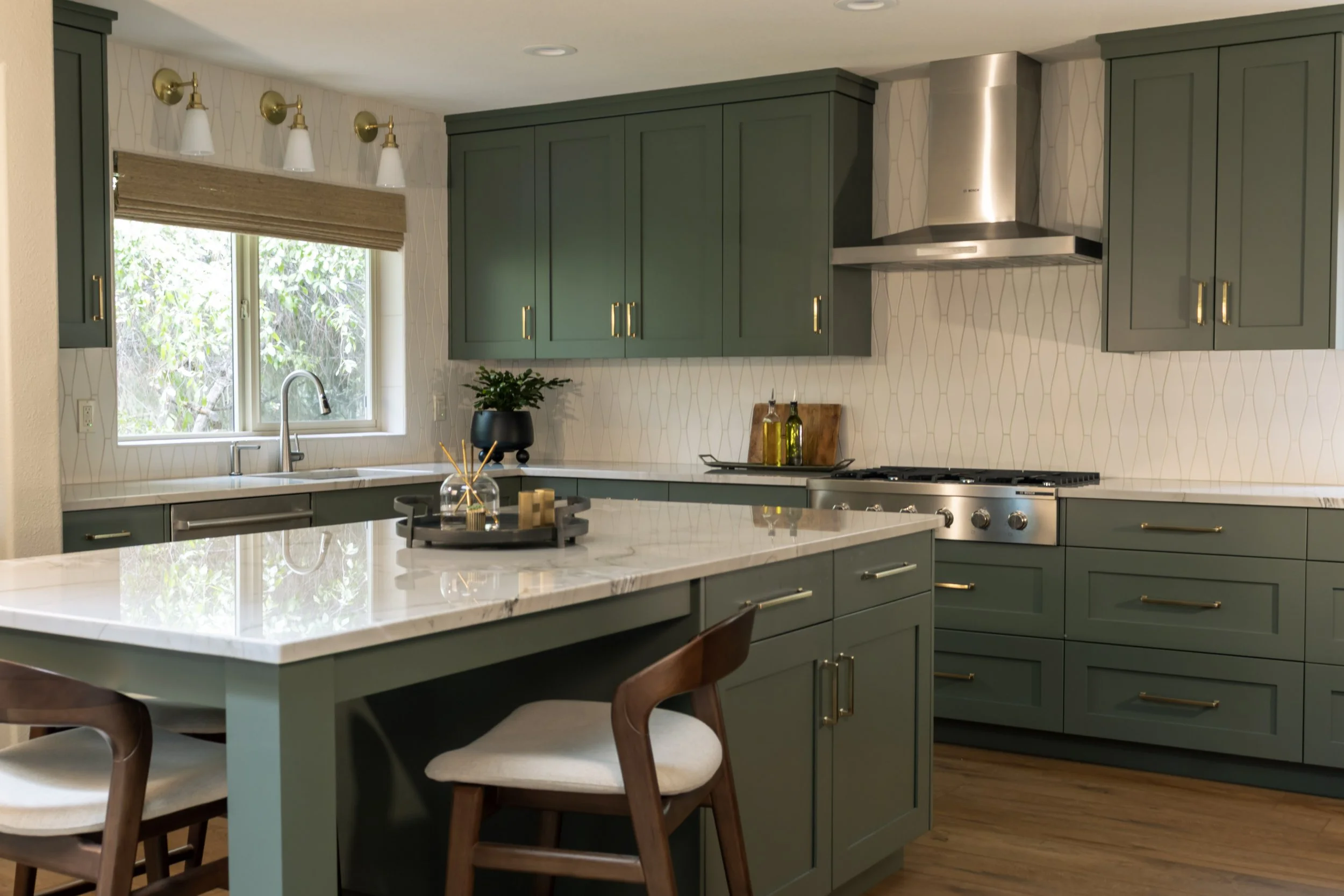 A modern kitchen with green cabinets, a white countertop island, and a window with a beige Roman shade. There are gold hardware handles, a stainless steel range hood, and a small potted plant on the counter.