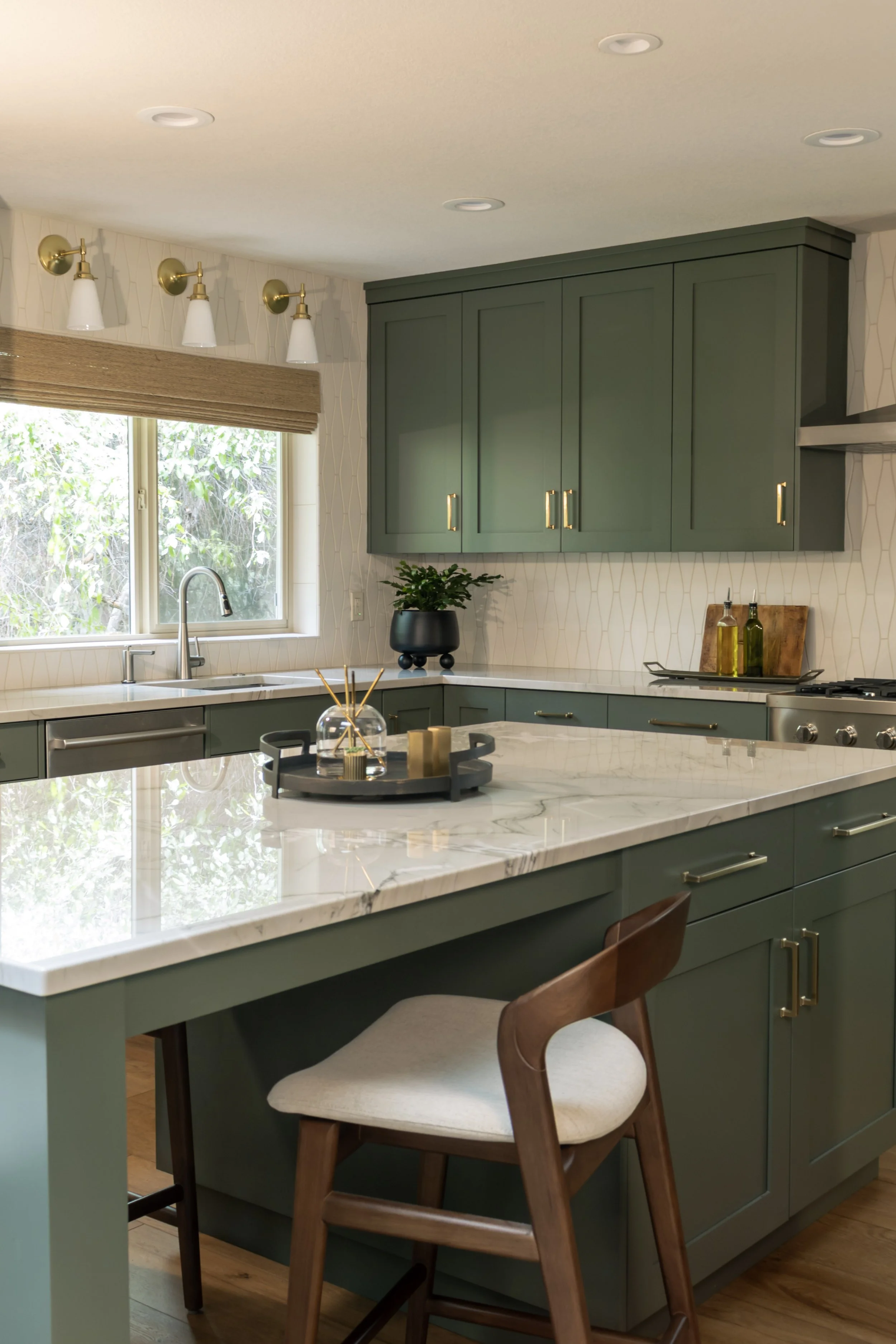 A modern kitchen with green cabinets and white marble countertop. There is a window with a beige roman shade, a black plant pot, and decorative oil bottles on the counter. Wooden chair with a cushioned seat is at the kitchen island.