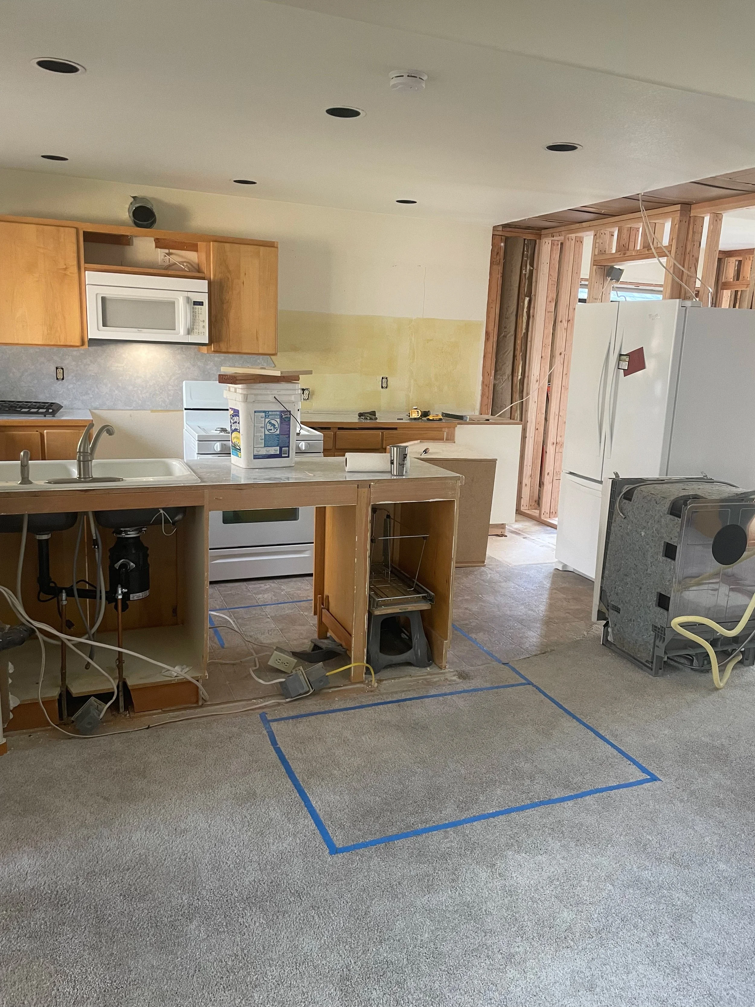 Room under construction with partially finished kitchen area, exposed wooden framing, and a marked section on the carpet outlined with blue painter's tape.