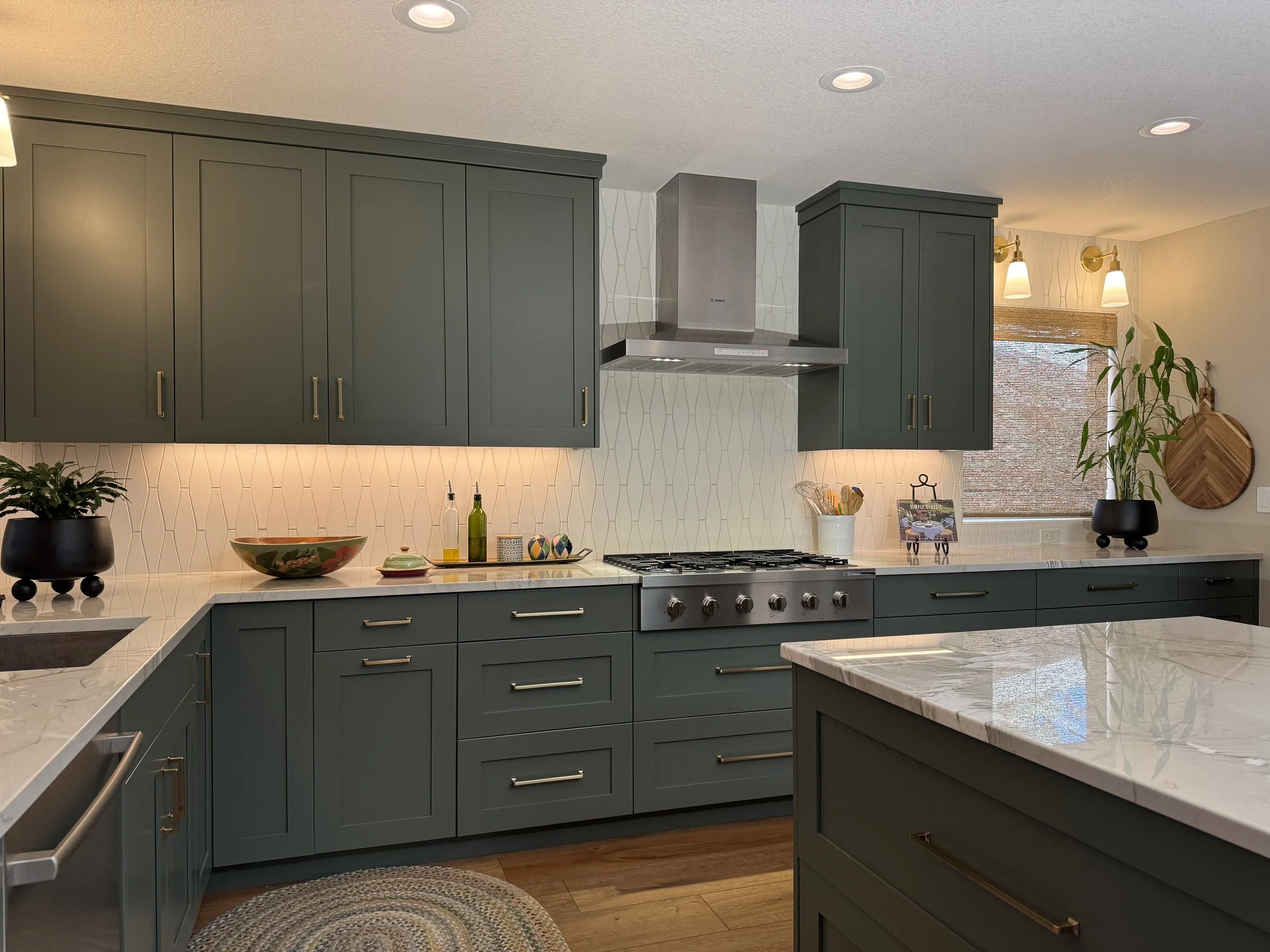 A modern kitchen with teal colored cabinets, white marble countertops, stainless steel range hood, and a window with a woven blind, decorated with potted plants, bowls, and bottles on the counter.