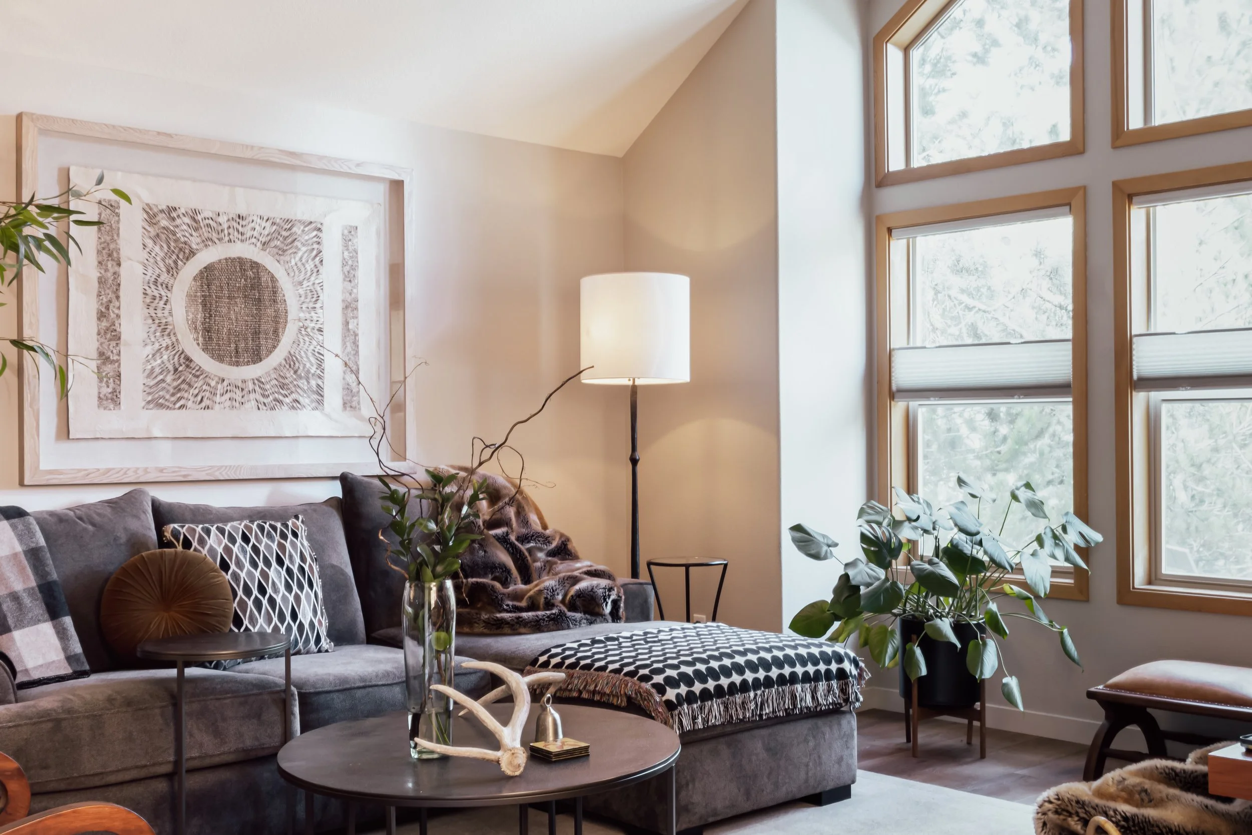 Living room with gray sofa, decorative pillows, potted plants, a large abstract black-and-white wall art, a white floor lamp, and large windows with wooden frames letting in natural light.