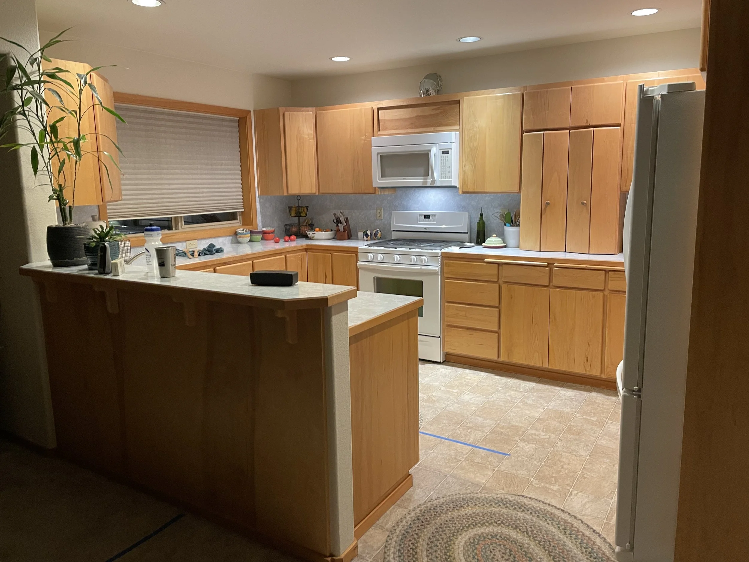 A kitchen with wooden cabinets, a white microwave, white stove, and various kitchen items on the counter, viewed from a doorway.