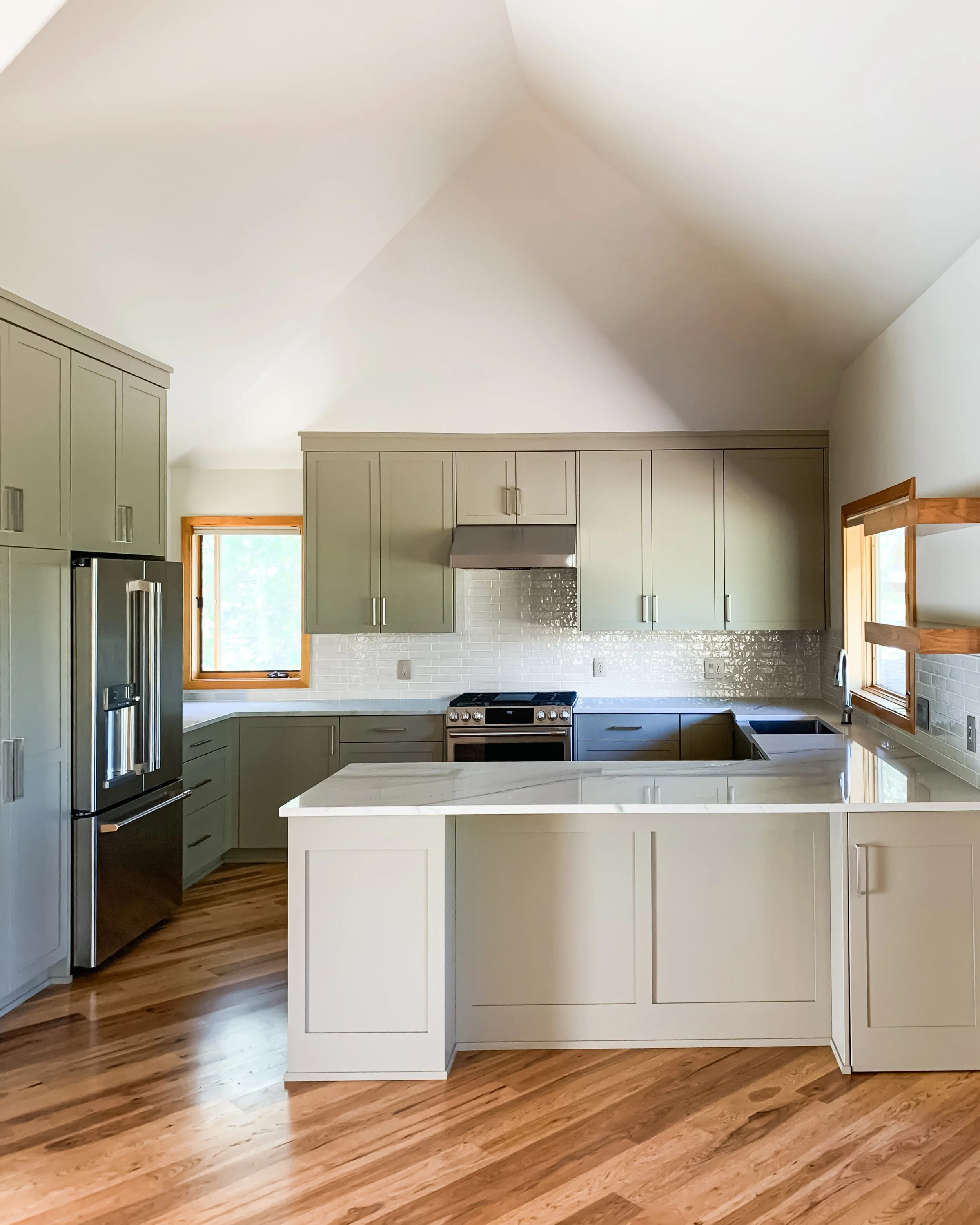 A modern kitchen featuring light grey cabinets, a center island with a white marble countertop, wooden flooring, a stainless steel refrigerator, a range with a vent hood, and two windows allowing natural light.