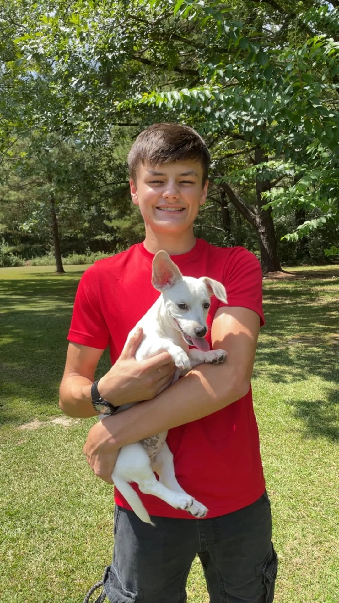A young man wearing a red shirt and black shorts is outdoors holding a white puppy with large ears, surrounded by green trees and grass on a sunny day.