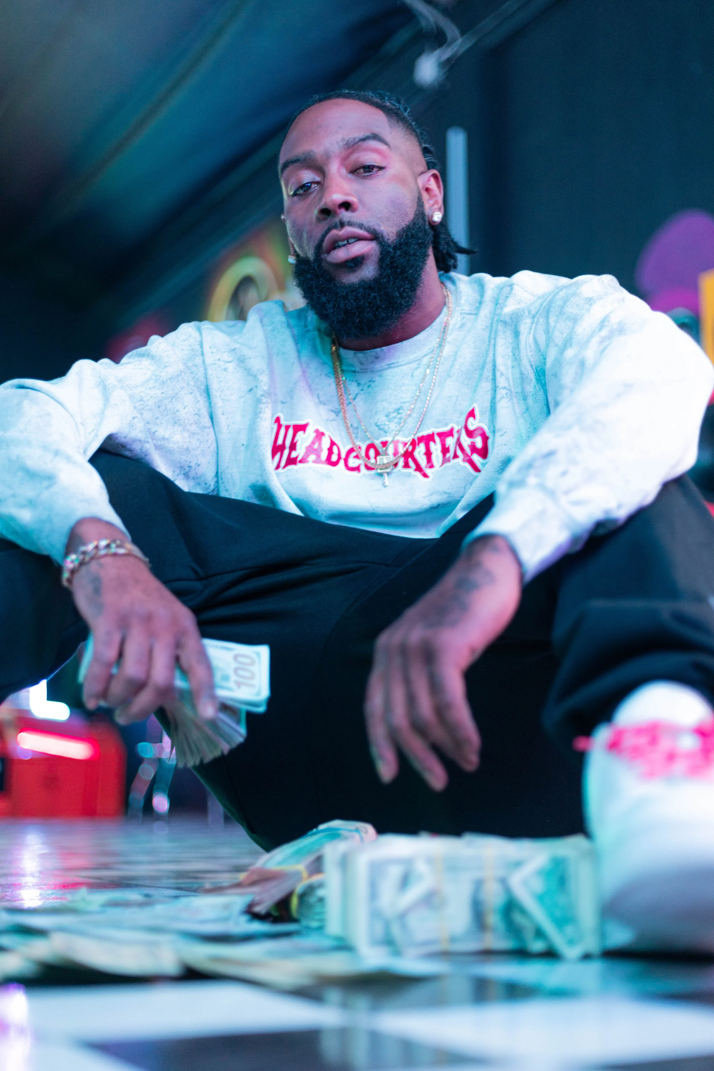 A man with a beard and jewelry, sitting on the floor, holding a stack of cash, with more cash spread out in front of him, in a colorful indoor setting.