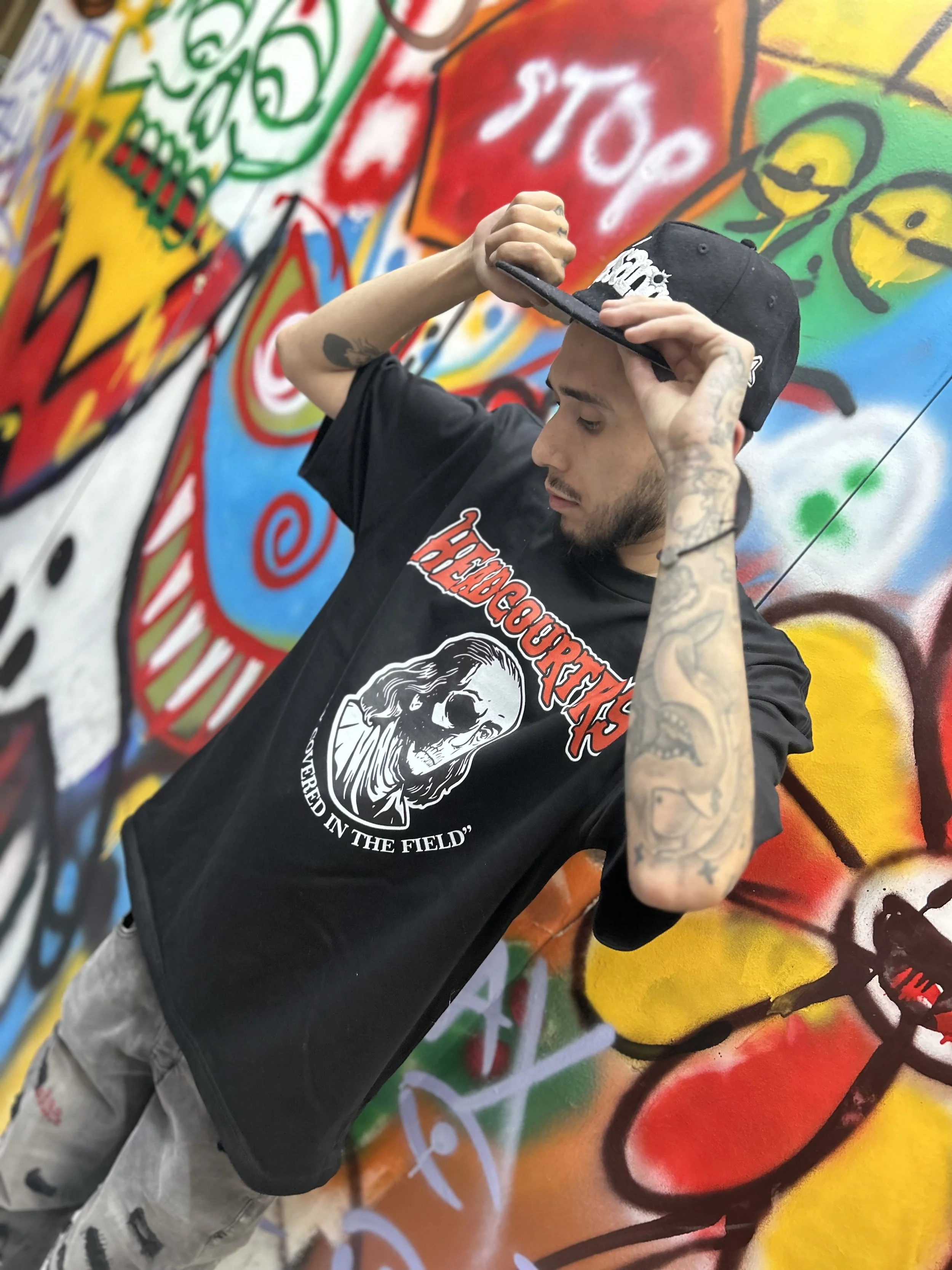 Young man with tattoos on his arms, wearing a black graphic “HEADCOURTERS” T-shirt and a black cap, stands in front of a colorful graffiti wall, adjusting his cap.