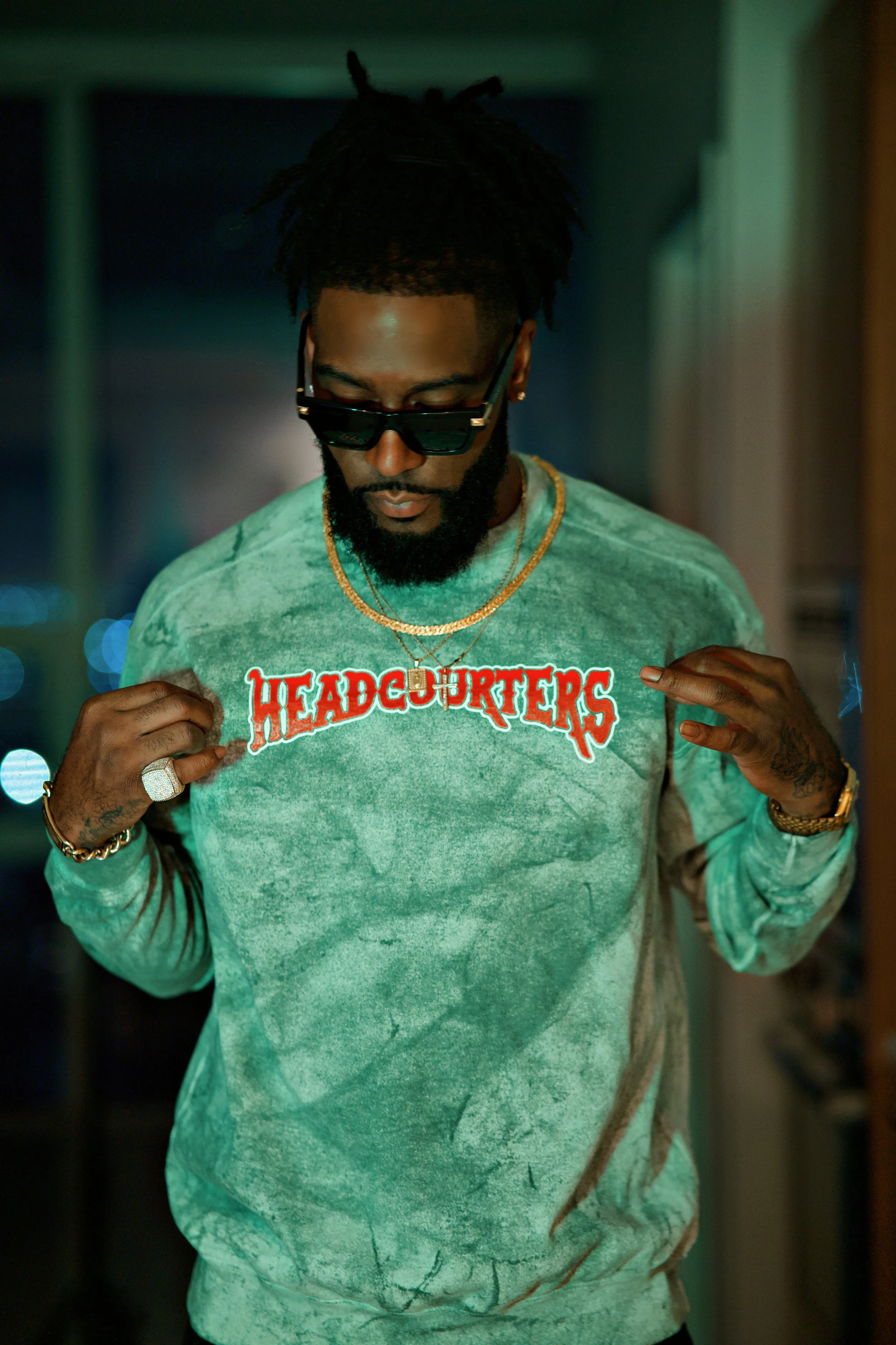 Man with dreadlocks, beard, and sunglasses wearing a green tie-dye sweatshirt that says 'HEADCOURTERS ' in red lettering, holding the sweater outwards with both hands.