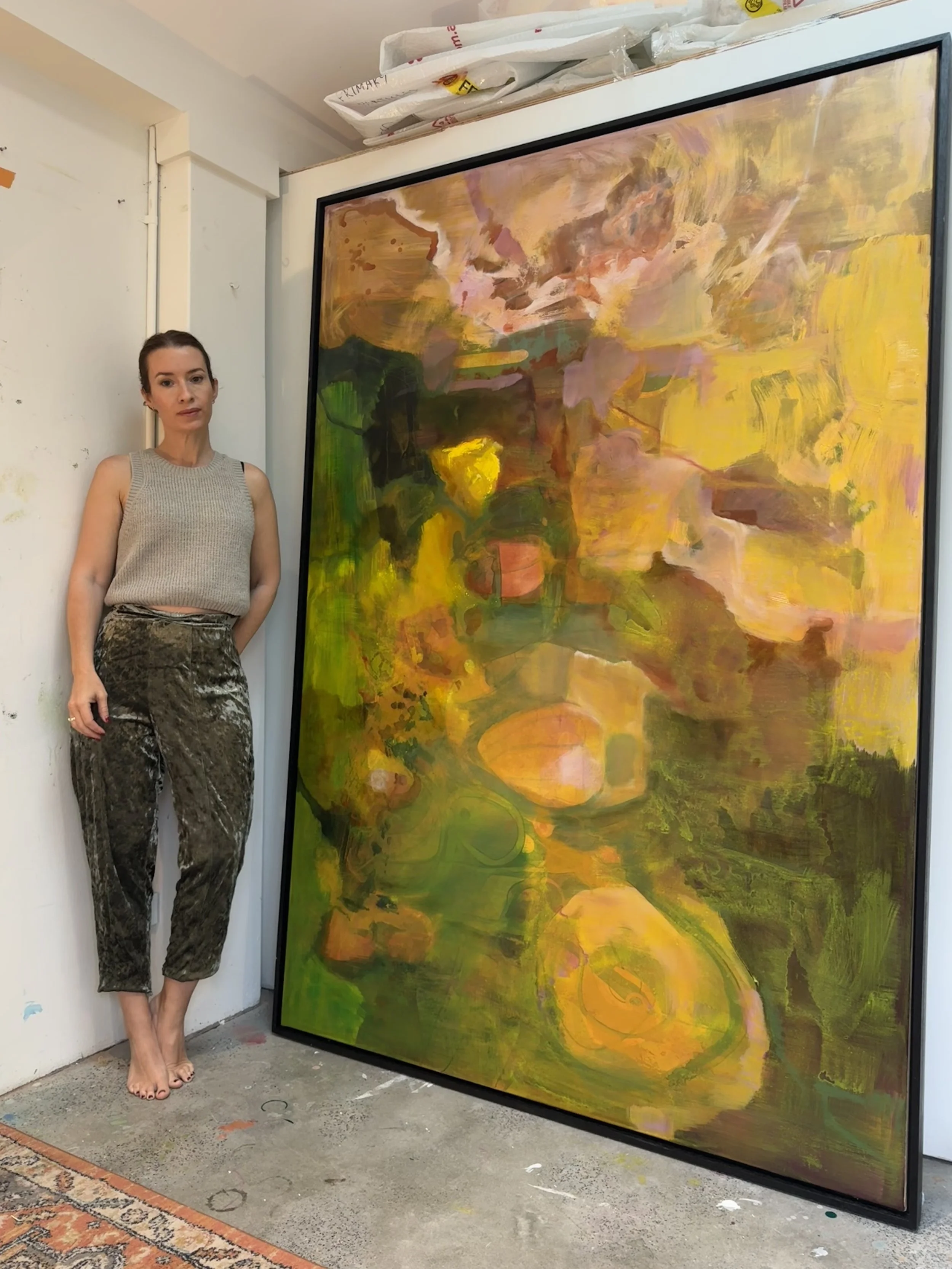 A woman stands beside a large colorful abstract painting in an art studio.