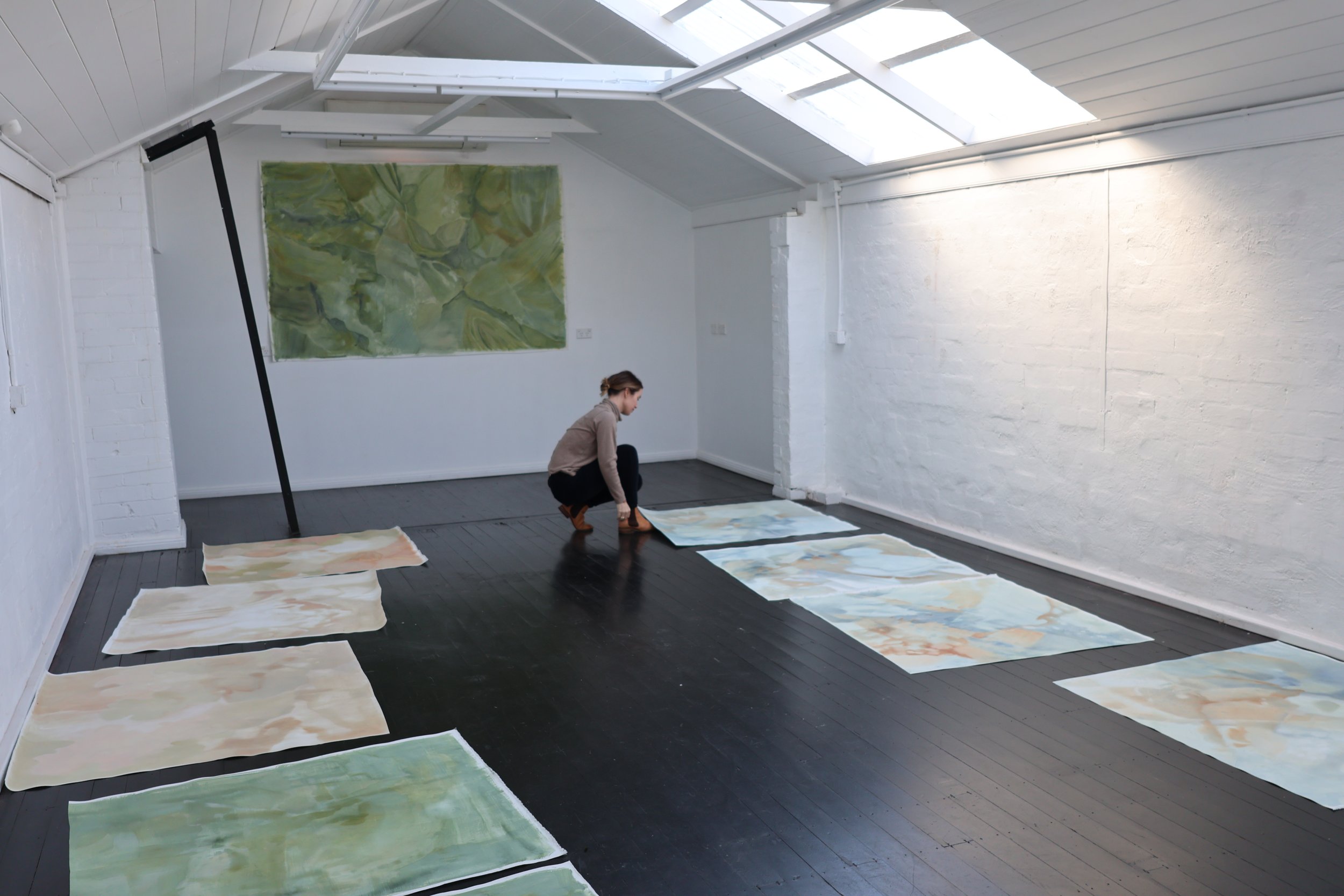 A woman in a grey top and black pants is kneeling on a black wooden floor, arranging large abstract paintings laid out on the floor, with a white brick wall and large windows above.