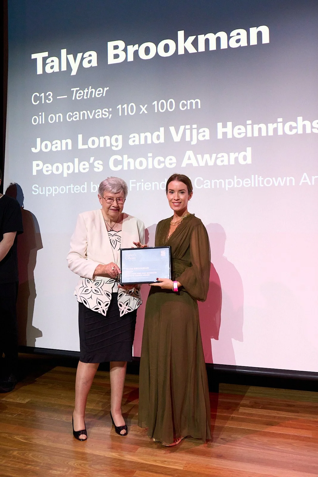 Two women standing on a stage, holding an award plaque, at an event honoring Talya Brookman for Joan Long and Vija Heinrichs People's Choice Award, with a large screen behind them displaying event details.