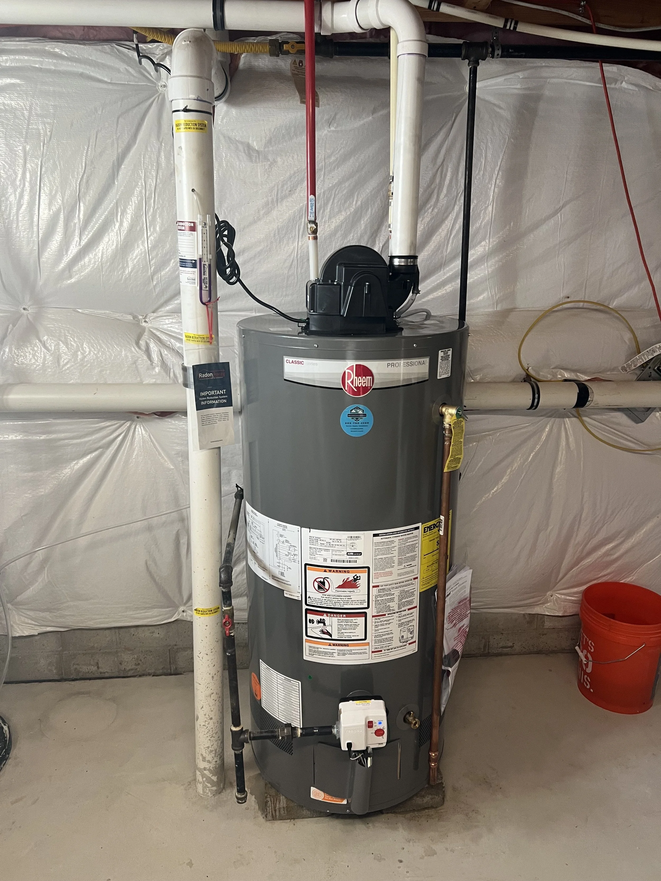 A water heater with copper pipes and a beige expansion tank attached, mounted on a white wall with visible cracks.