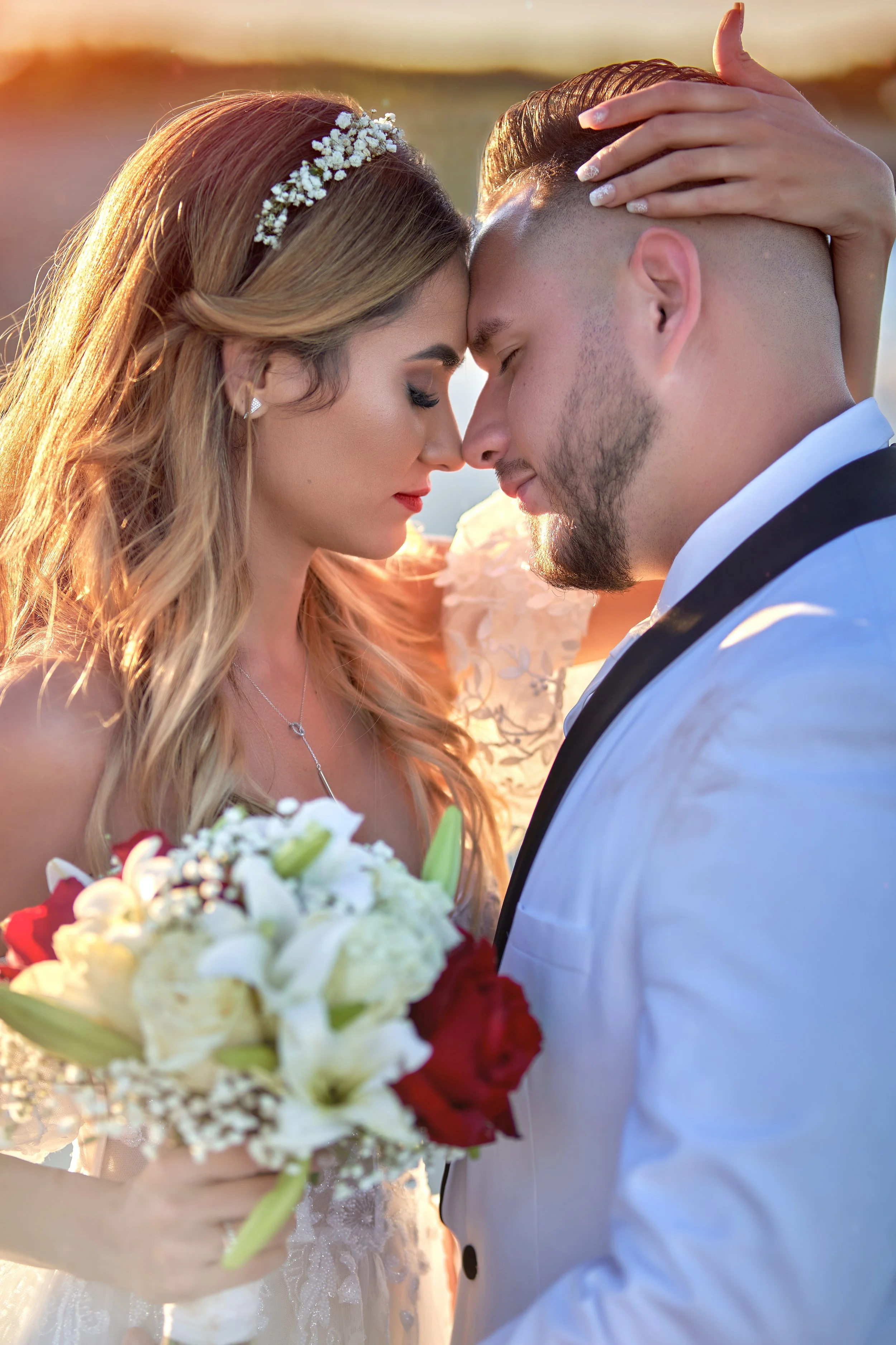 Novios en su boda, con la frente tocándose, ella con ramo de flores, al atardecer.