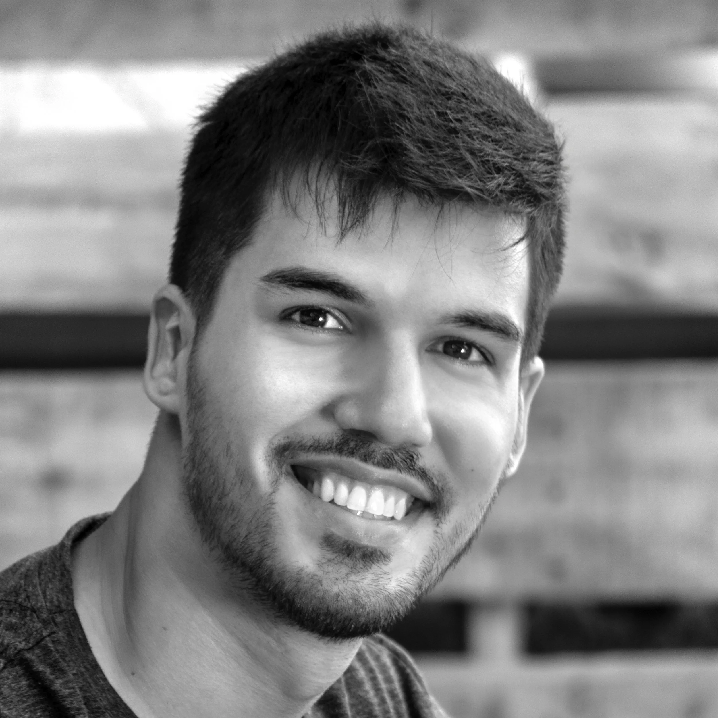 Retrato en primer plano de un joven sonriente con barba y cabello oscuro, fondo difuso de lo que parece una estructura de madera.