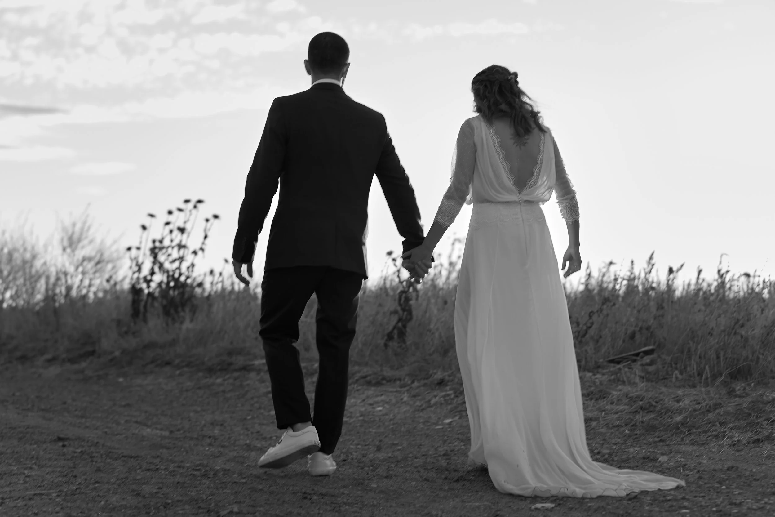 Pareja caminando al atardecer en un campo, ella con vestido de novia y él con traje. La imagen en blanco y negro.