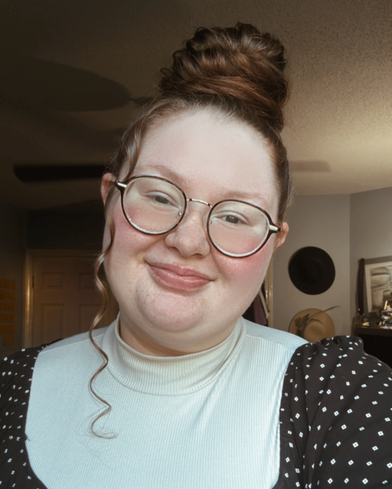 A young woman with glasses, red hair in a top bun, smiling indoors with a beige and gray background, wearing a light-colored turtleneck with black polka-dotted sleeves.