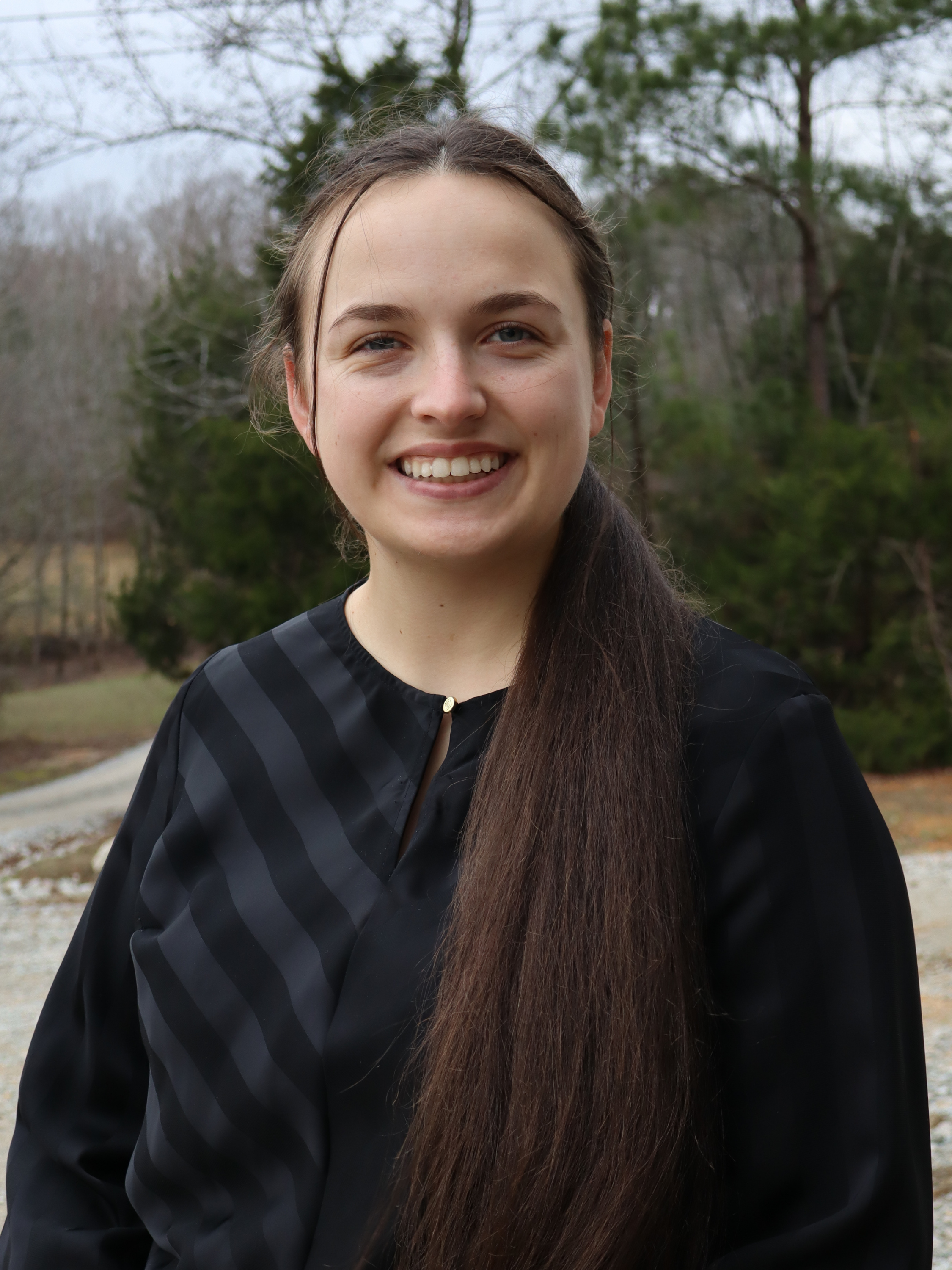 A young woman with long dark hair, smiling, outdoors in a natural setting with trees in the background.