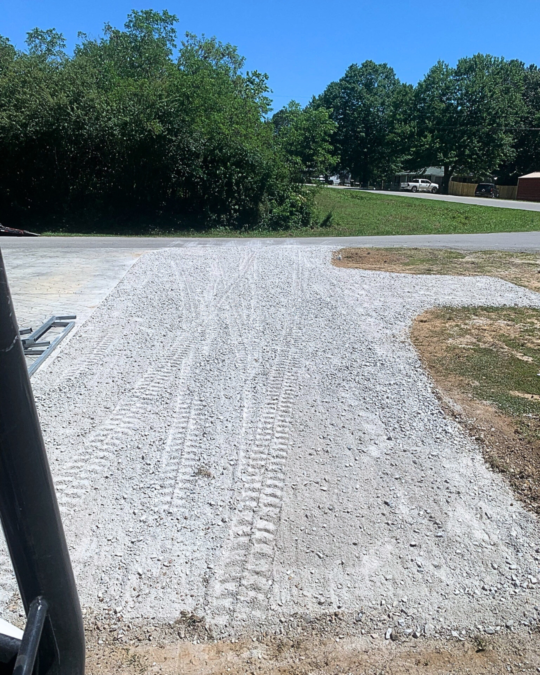 Gravel driveway with visible tire tracks leading to a paved road, with trees and parked cars in the background under a clear blue sky.