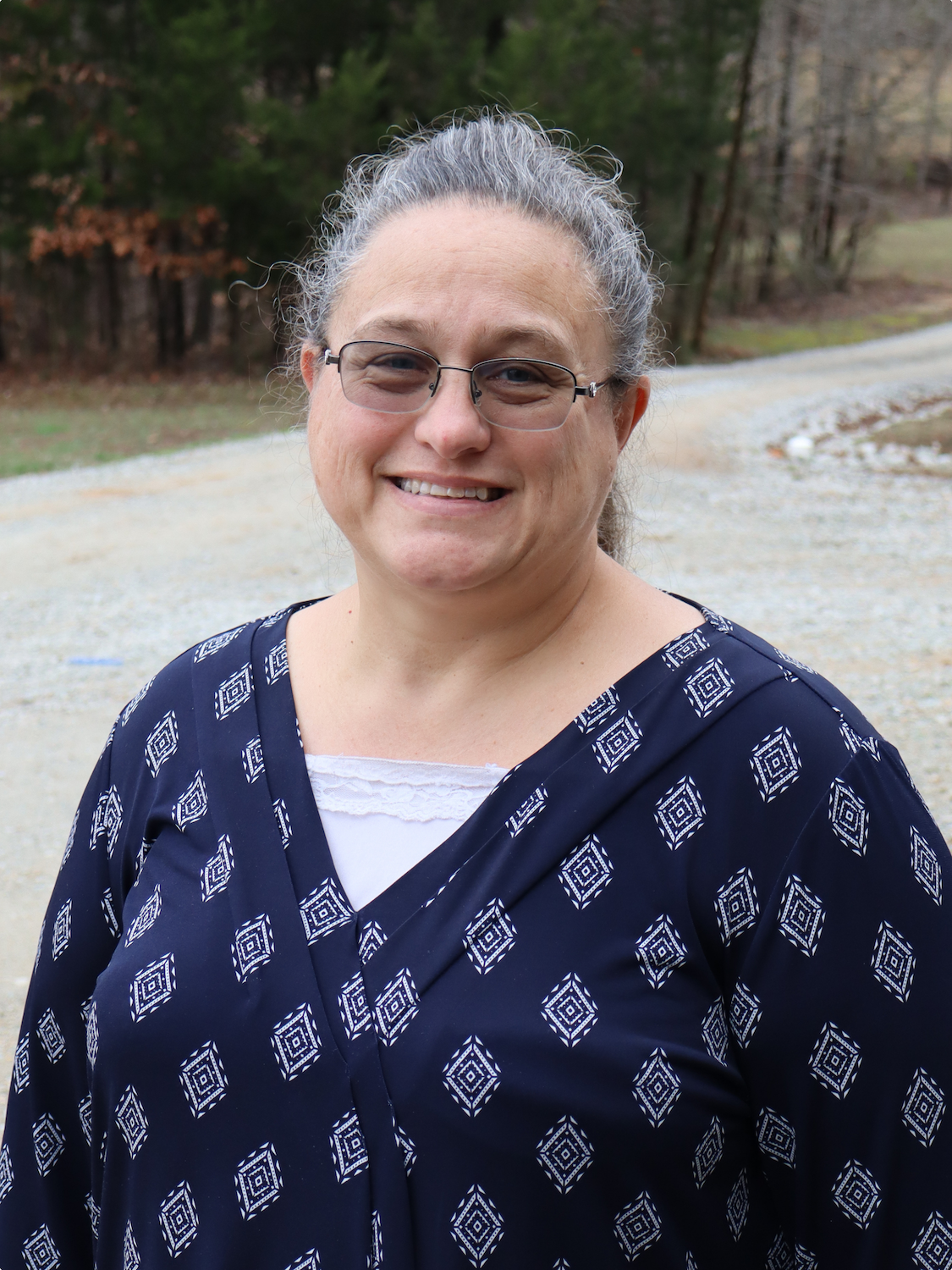 A woman with gray hair pulled back, wearing glasses, a navy blue patterned blouse, and a white top underneath, standing outdoors on a gravel path with trees in the background.
