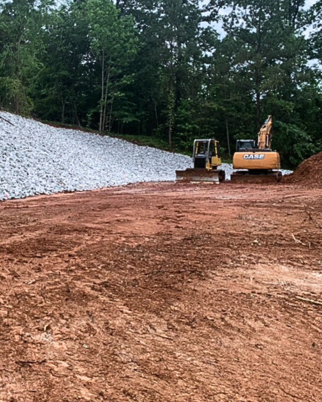 Construction site with two compact track loaders moving dirt, surrounded by trees and a gravel embankment.